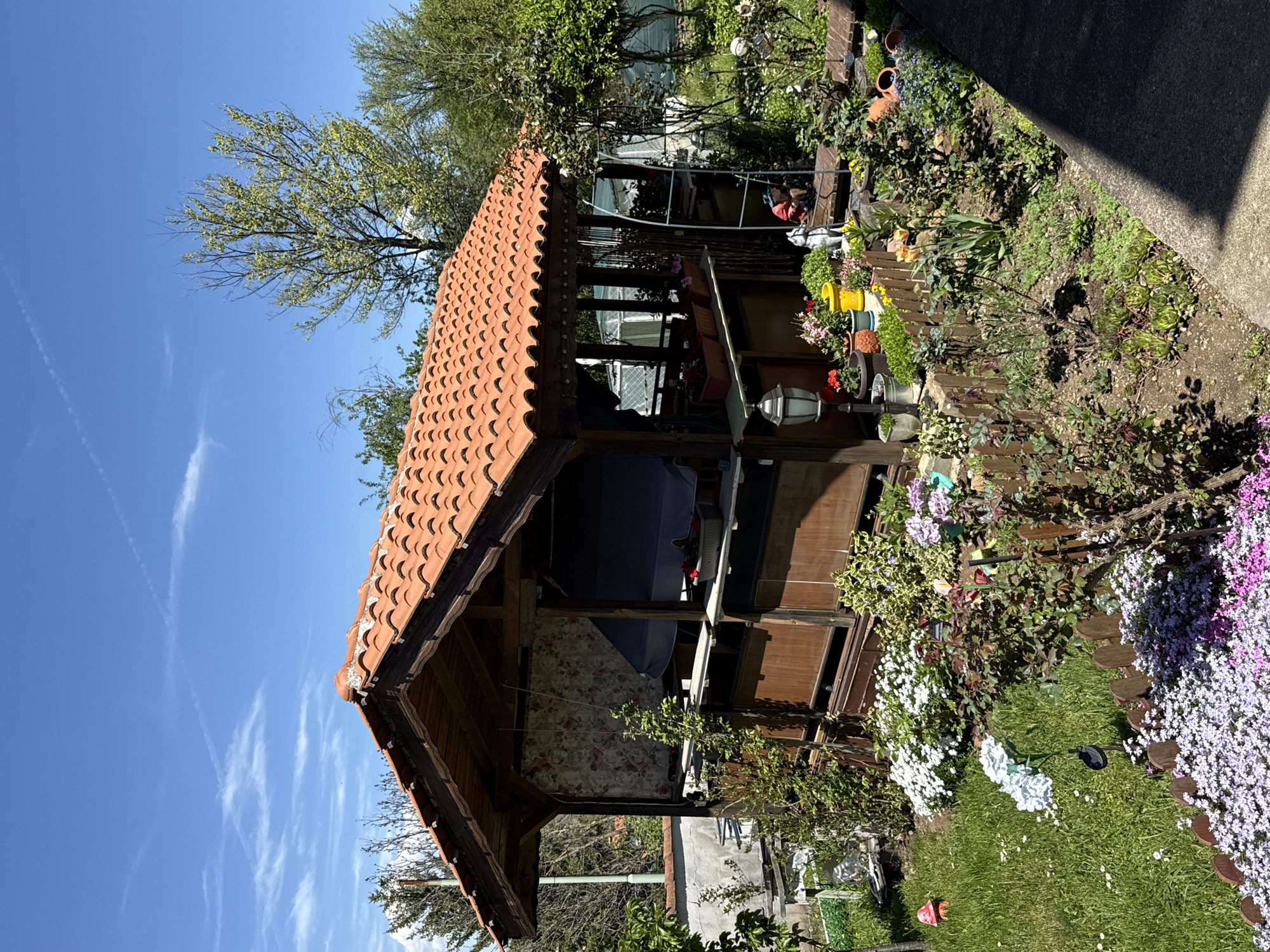 The image shows a wooden gazebo with a red tile roof surrounded by a garden in Selishten Dol, Bulgaria. The gazebo is nestled among lush greenery and colorful flowers. The sky is blue with wispy clouds. The garden features a variety of plants and flowers, including white and purple blooms. A fence borders the garden. A portion of a nearby building is visible.