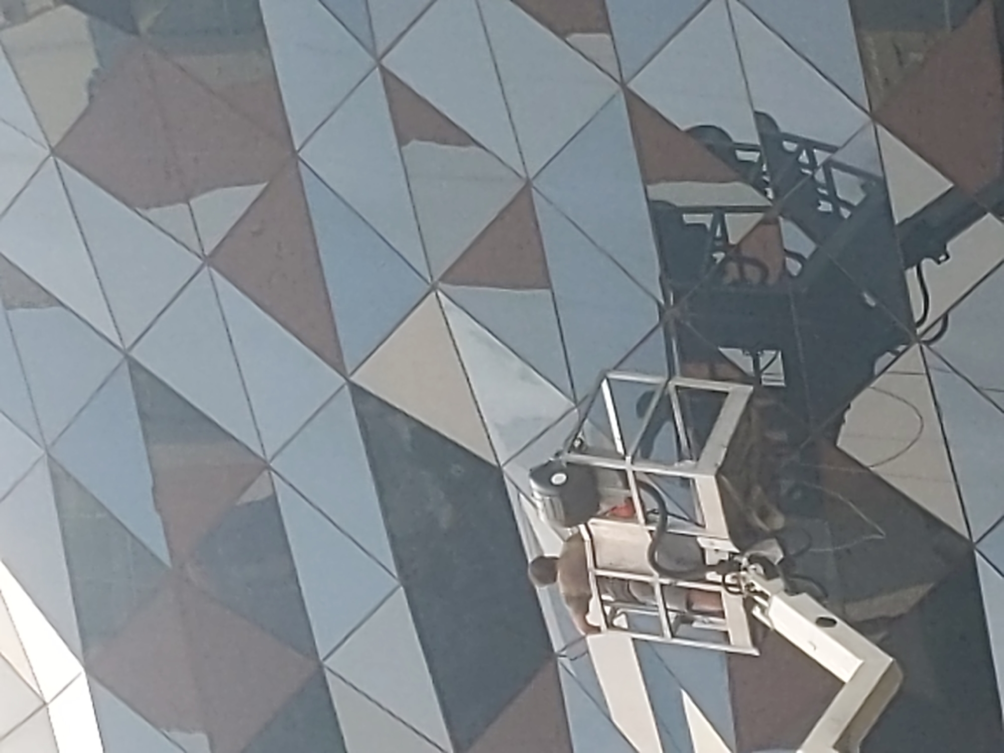 A person stands in a cherry picker, performing work on the exterior of a building. The building's facade is composed of triangular panels in varying shades of blue, grey, and beige, creating a geometric pattern. The cherry picker is white and metallic, with a platform where the person stands. The person is wearing a light-colored shirt and shorts, and their back is to the camera. The building appears to be modern, with a reflective surface that mirrors the sky and surrounding environment. The image was taken in Kyiv, Ukraine.