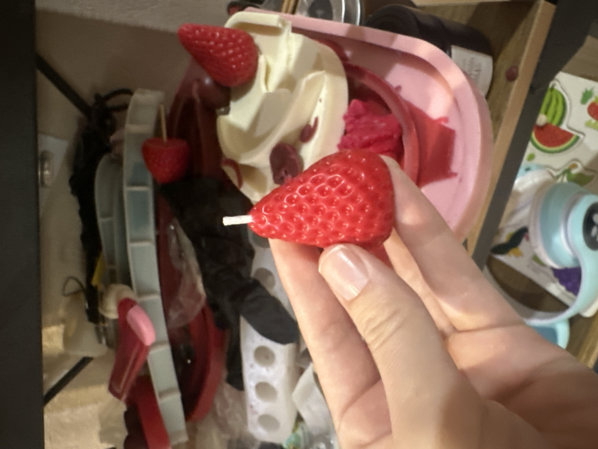 A close-up shot shows a hand holding a red, strawberry-shaped candle. The candle is detailed with small indentations to mimic the texture of a strawberry. The background is cluttered with various items, including what appears to be a pink dish filled with white and red material that resembles dessert or ice cream, and other red and clear plastic containers. The scene is dimly lit, suggesting an indoor setting. The image does not provide any information about the location in Vladivostok, Russia.