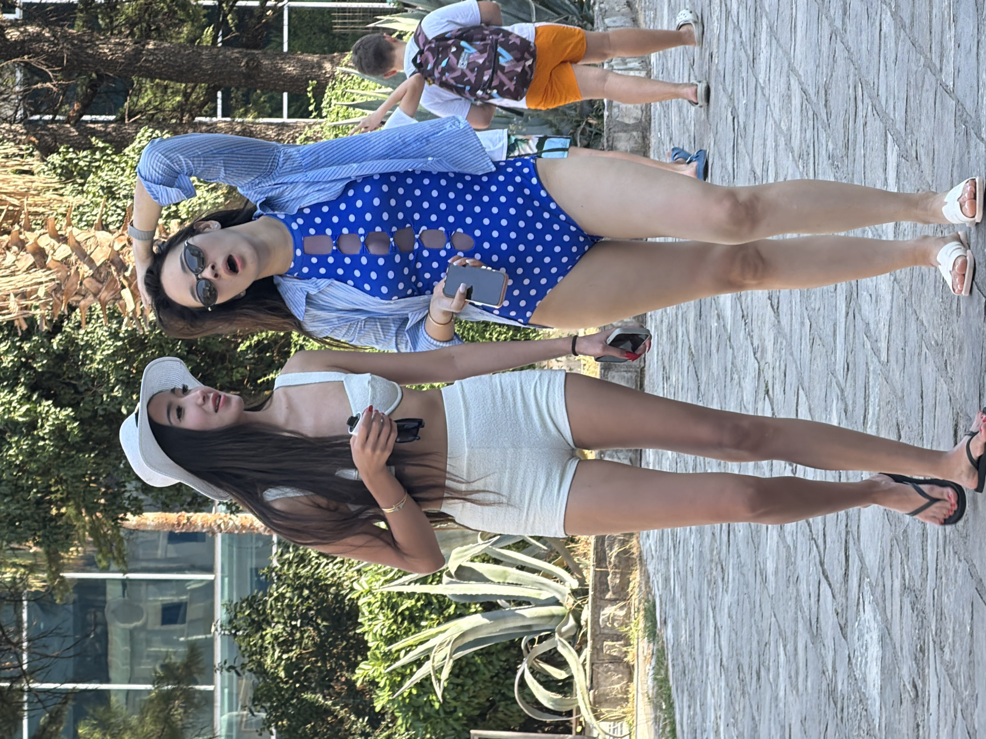 Two young women are walking on a paved walkway in Budva, Montenegro. The woman on the left is wearing a white sun hat, a white bikini top, and white shorts. She is holding a dark object in her right hand and a cell phone in her left hand. The woman on the right is wearing sunglasses, a blue and white polka dot swimsuit, and a blue striped shirt. She is holding her right hand to her head and a cell phone in her left hand. In the background, a young boy wearing a backpack and orange shorts is walking away from the camera. Lush green vegetation and trees surround the walkway. The ground is paved with stone tiles.