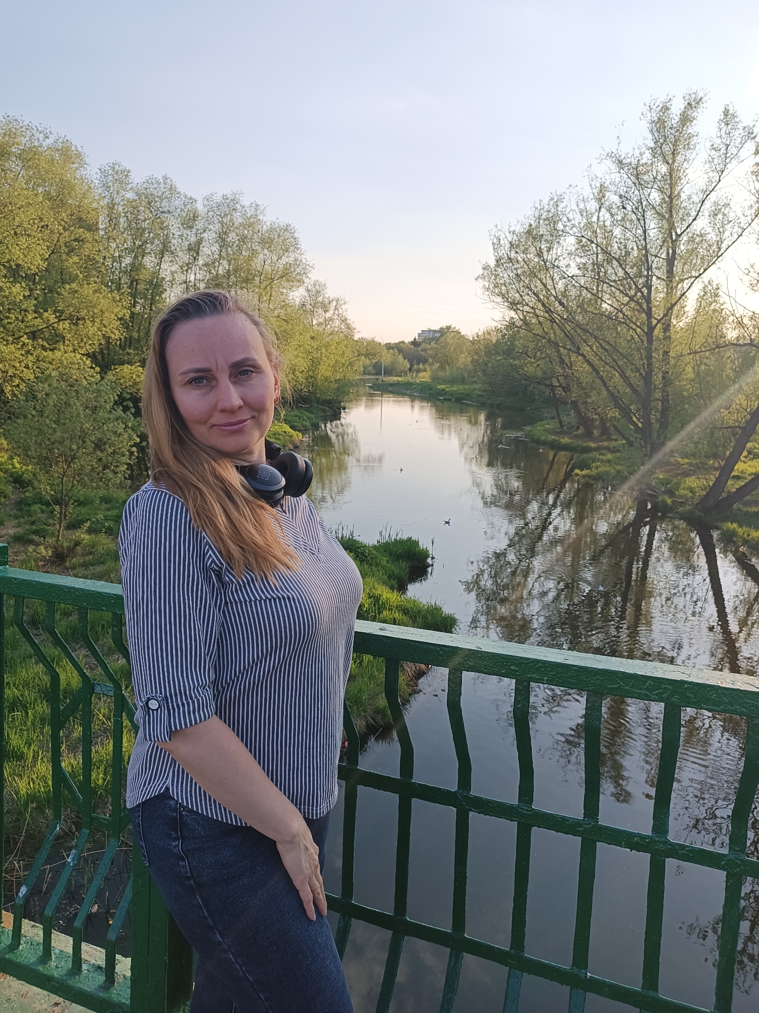 A medium shot shows a woman standing on a green metal bridge with a river and trees in the background. She is fair-skinned with long auburn hair and is wearing a striped shirt and dark jeans. She has black headphones around her neck. The river behind her reflects the surrounding trees. The environment is bright, suggesting daytime. The exact location in the city of Unknown, Unknown is not discernible from the image.