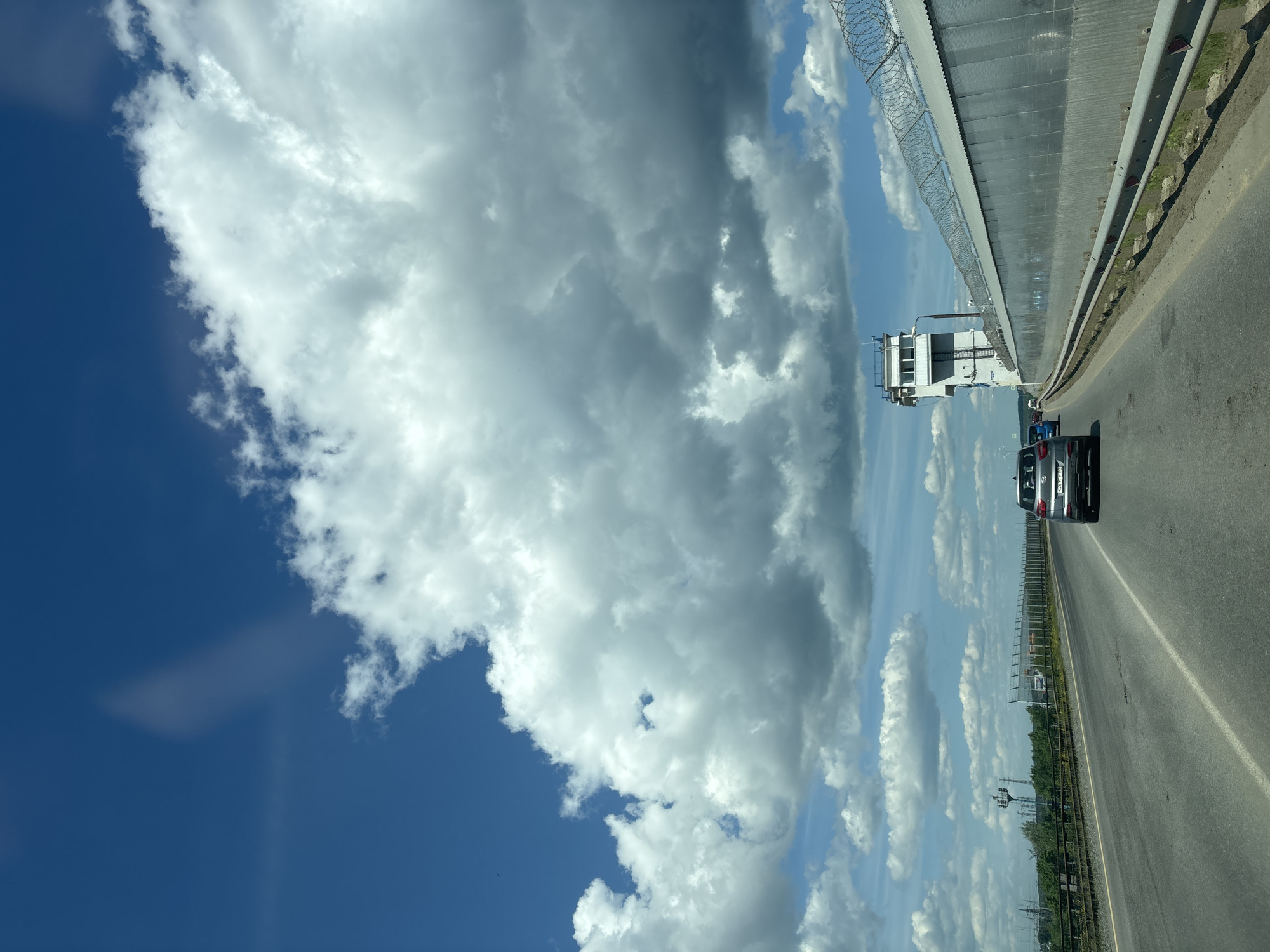 The image captures a daytime scene from a vehicle traveling on a multi-lane asphalt road under a partly cloudy sky near Chaikovsky, Russia. Large, predominantly white and grey cumulus clouds dominate the upper and mid-portions of the sky, with patches of clear deep blue sky visible in the upper left. A faint, elongated streak of light or reflection is present in the upper left sky.
The road, appearing to be a bridge or elevated highway section, shows signs of wear and patching. White lane markings, including a solid line for the shoulder and a broken line for the main lane, are visible. Metal guardrails run along both sides of the roadway.
In the immediate right foreground, a gray Hyundai Creta SUV, bearing the Russian license plate "X267FF 759", is seen driving away from the viewer. Further ahead, other vehicles, including a blue car, are visible on the road.
To the right of the road, a long, tall wall, constructed from corrugated metal panels, extends into the distance. This wall, approximately three to four meters in height, is topped with multiple coils of razor wire, indicating a secure or restricted area. Behind this wall and slightly to the right, a prominent multi-story, light-colored concrete building stands tall. This structure features small, rectangular windows and an external staircase or ladder, characteristic of a control tower or observation post. This specific building and wall are part of the infrastructure associated with the Votkinsk Hydroelectric Power Station dam, located in the city of Chaikovsky, Perm Krai, Russia.
To the left of the road, beyond the guardrail, a fence or railing is visible, followed by a dense line of green deciduous trees. In the middle distance on the left, several tall utility poles or transmission towers can be seen, suggesting an industrial or power infrastructure presence. The overall environment is one of an open highway under bright daylight.