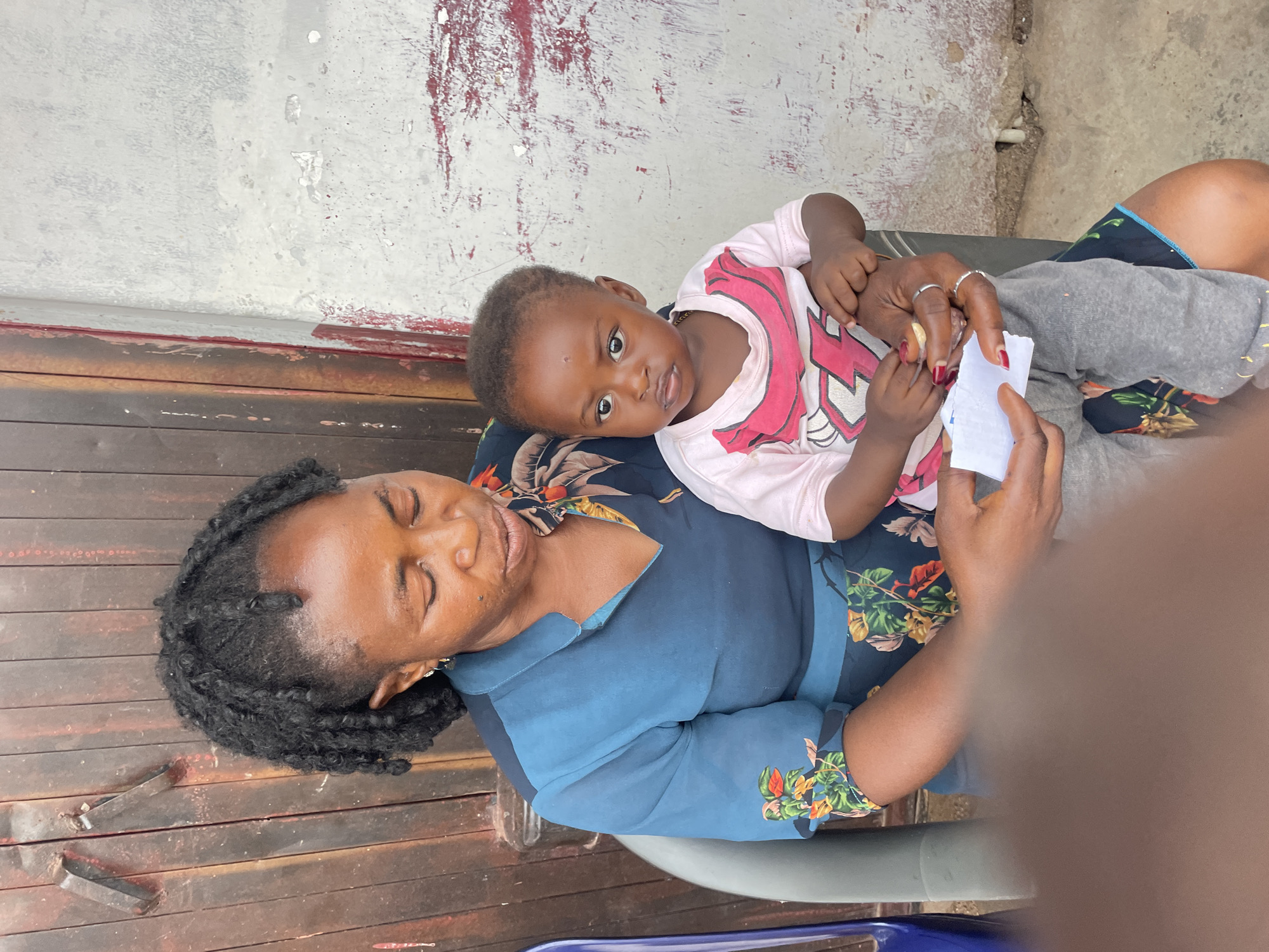 The image shows an adult woman sitting outside with a young child resting on her lap. The woman has dark skin, and her hair is styled in braids or twists. She is wearing a blue floral-patterned top. The child is also dark-skinned and wearing a light pink shirt with a cartoon-like design. The woman is holding a small white card in her hands, and the child is reaching for it.
The background consists of a weathered wall with peeling paint and a wooden surface. The setting is presumed to be Jos, Nigeria, based on the prompt's instructions. There is an object that could be a chair partly visible under the woman, which suggests they are seated.