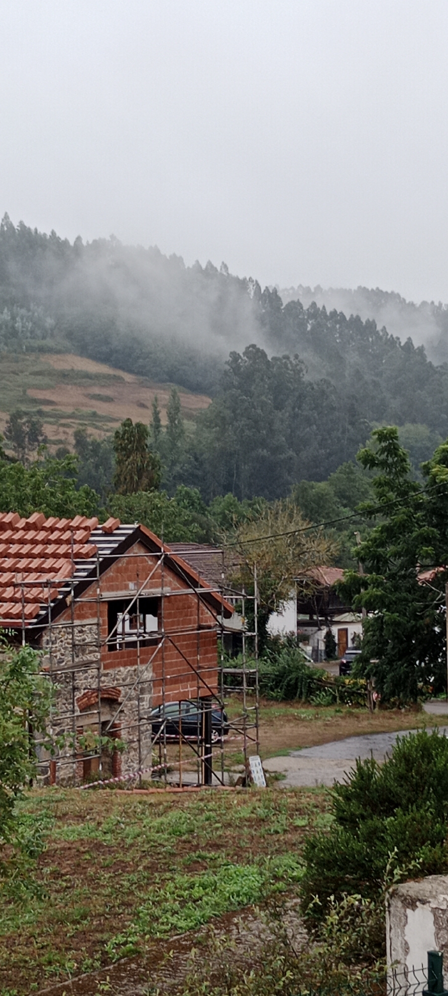 The image shows a rural scene in the municipality of Pravia, Spain, characterized by a cloudy and misty sky and lush green vegetation covering rolling hills. In the foreground, a house is under construction, with exposed brickwork and scaffolding surrounding it. The roof is partially tiled with red ceramic tiles. A dark-colored car is parked near the construction site. In the background, a dense forest covers the hills, with patches of bare earth visible on some slopes. The overall atmosphere is damp and overcast, suggesting a recent or ongoing rainfall.