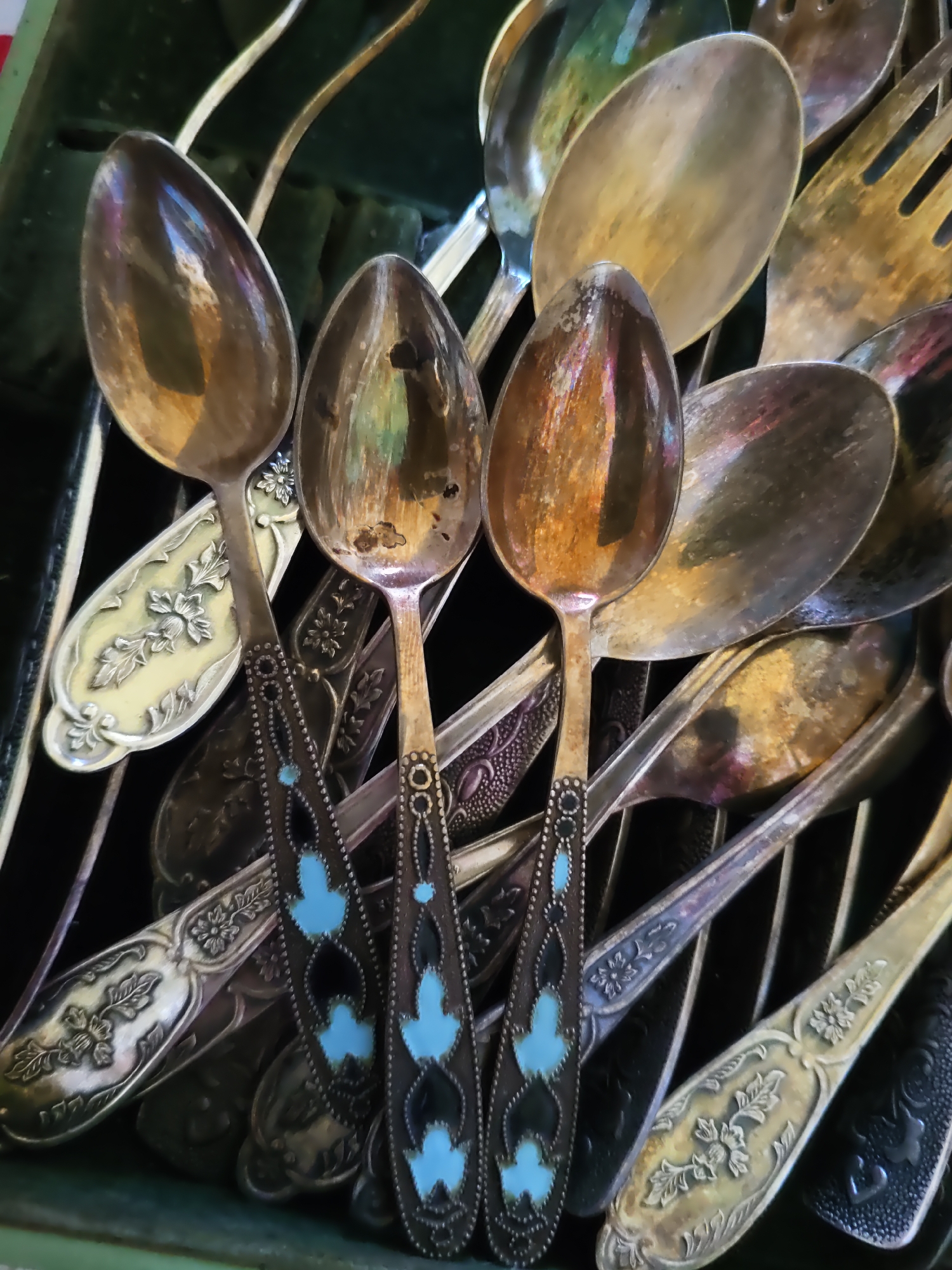 A close-up, overhead shot shows a jumbled collection of vintage silverware, primarily spoons and some forks, piled in a green container. The spoons vary in design; some have ornate floral patterns etched onto their handles and are tarnished with age, displaying a warm, golden hue mixed with darker patina. Others feature darker handles with inlaid turquoise and black designs, creating a contrasting, decorative element. The reflective surfaces of the silverware capture the surrounding light, creating colorful glints and distorted reflections. The overall impression is one of a disorganized yet aesthetically rich collection of antique utensils. There is no information in the image to suggest a specific location in Rostov-on-Don, Russia, or any discernible actions or interactions between people.