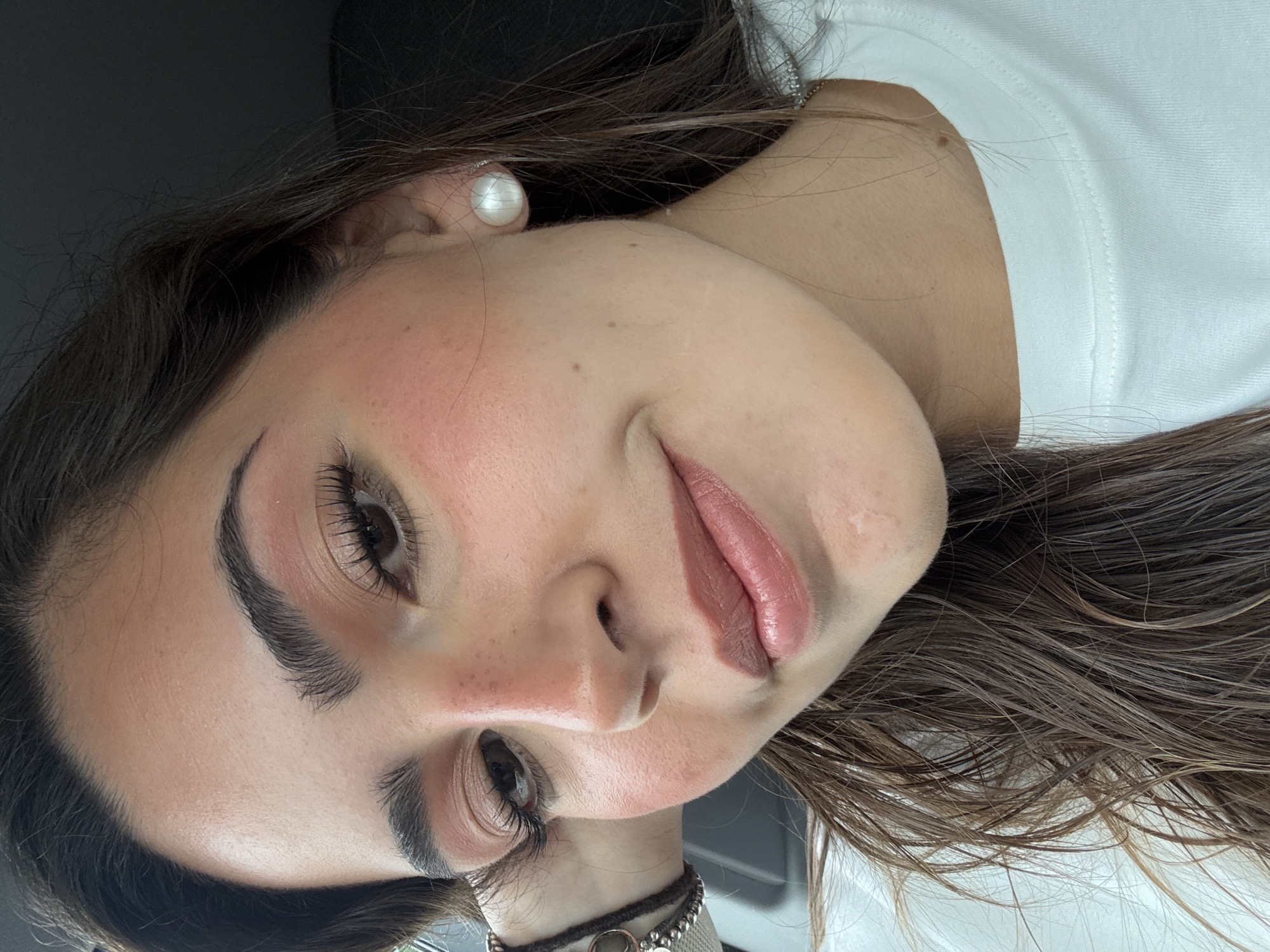 A close-up, slightly rotated portrait shows a light-skinned woman with brown hair and eyes. She has dark eyebrows and makeup applied to her eyelids, cheeks, and lips. She wears a pearl earring in her left ear and a bracelet on her left wrist. She is wearing a white shirt. The background is indistinct but suggests an interior setting. The location within the city of San Antonio is not discernible from the image.