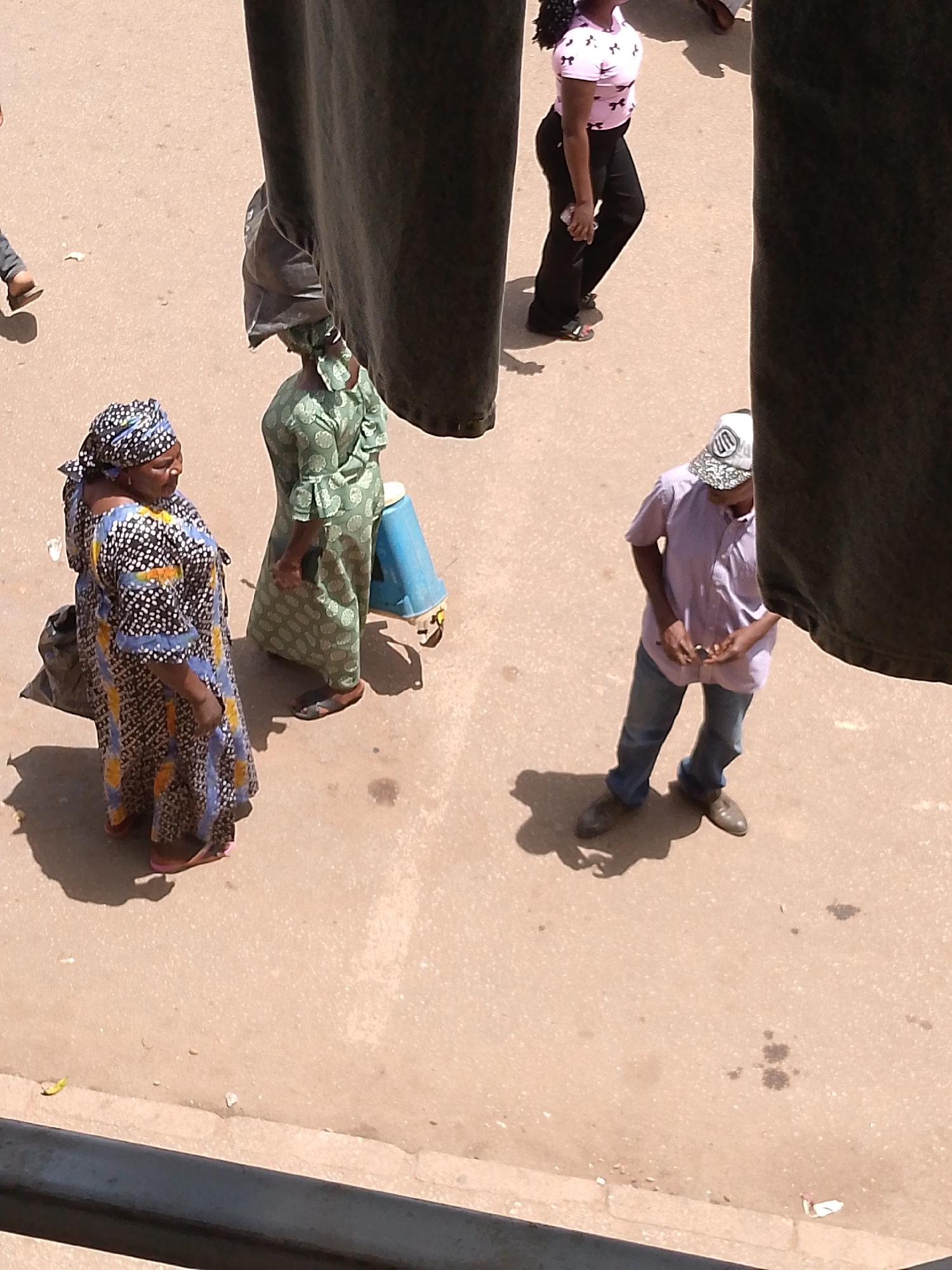 The image shows an outdoor scene in Bukuru, Nigeria, featuring several people on what appears to be a paved street or open area. From an elevated perspective, a woman in a colorful patterned dress stands on the left. Beside her is another woman in a green patterned dress with something on her head, and a blue container in her left hand. To the right of them, a man in a purple shirt, jeans, and a baseball cap looks down at something in his hands. Further in the background is a woman wearing a pink shirt and dark pants. The scene is partially obscured by dark fabric hanging in the foreground.