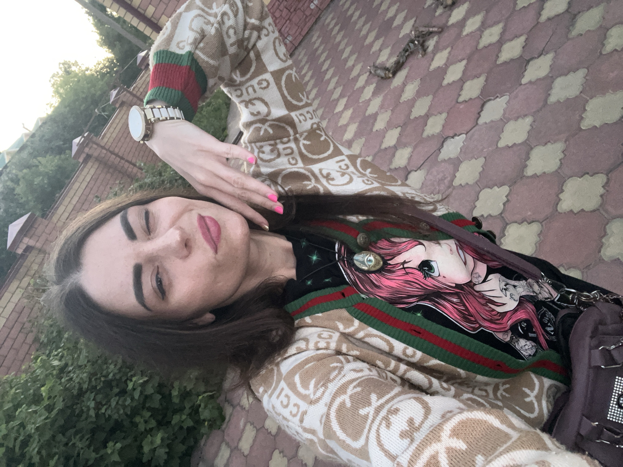 A medium-shot selfie of a woman in Krasnoe-na-Volge, Russia, shows her winking and posing outdoors. She is wearing a beige cardigan with a repeating GUCCl logo pattern and a black t-shirt featuring an anime-style graphic. She has a necklace with a large, circular pendant. A dark-colored purse with silver embellishments hangs from her left shoulder. The background consists of a brick fence and lush green foliage, with a patterned paved walkway visible in the foreground. The lighting suggests it is late afternoon or early evening.