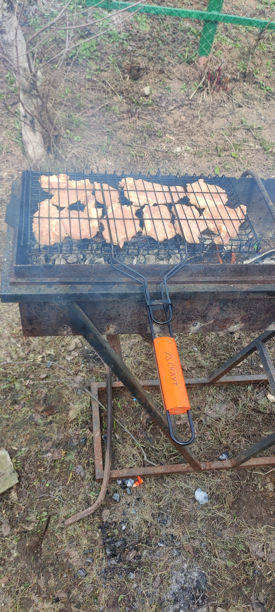 The image shows a metal grill filled with raw meat, likely pork, being cooked outdoors. The grill is placed on a rustic, possibly homemade, metal barbecue structure. An orange handle is visible, attached to the grill. The ground around the barbecue is covered with dry grass and some scattered debris, including what appears to be burnt charcoal. In the background, there is a fence with green posts and wire mesh, along with some trees and bushes. The location is Прикамский, Russia.