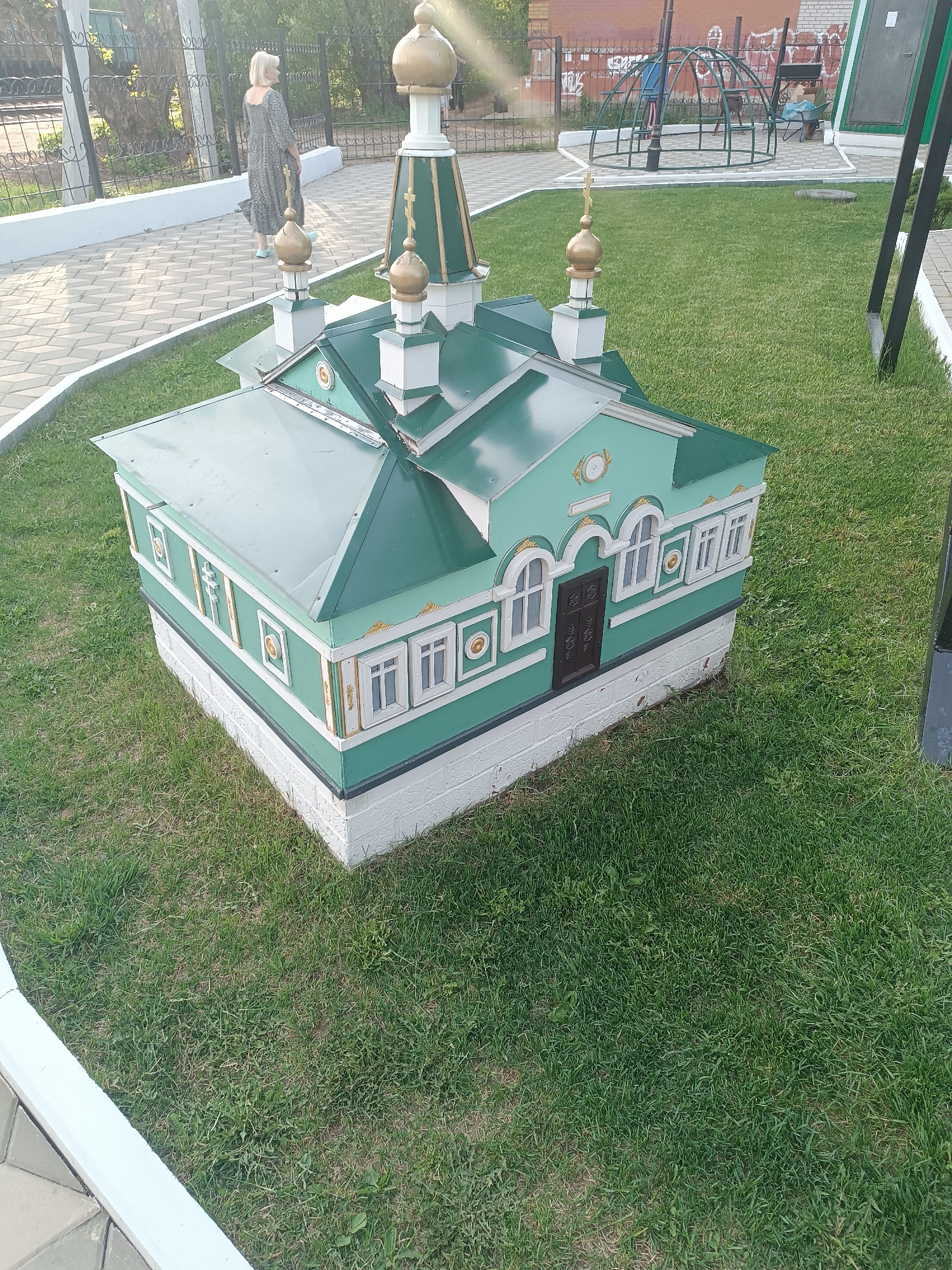 A green miniature building of a cathedral or church sits on green grass in an outdoor setting. The building is a square structure with a green roof and several golden domes on top. White trim borders the green walls and windows. A dark brown door is also visible. A woman in a long dress walks along a paved path in the background. A playground is in the distant background, behind the cathedral model.