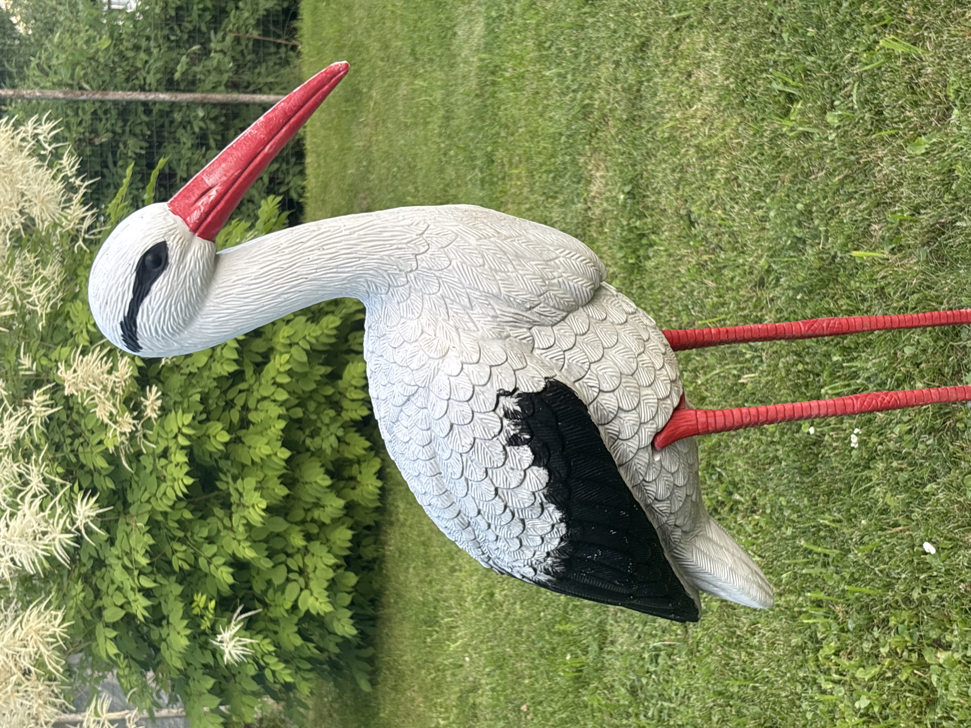 The image shows a decorative stork statue standing on a green lawn. The statue is white with black wings and a red beak and legs. It is positioned in the foreground, with a lush green bush and white flowers in the background. The lighting suggests it is daytime, likely outdoors in a garden or park setting. There is no indication of this location being in the city of Yermolino, Russia.