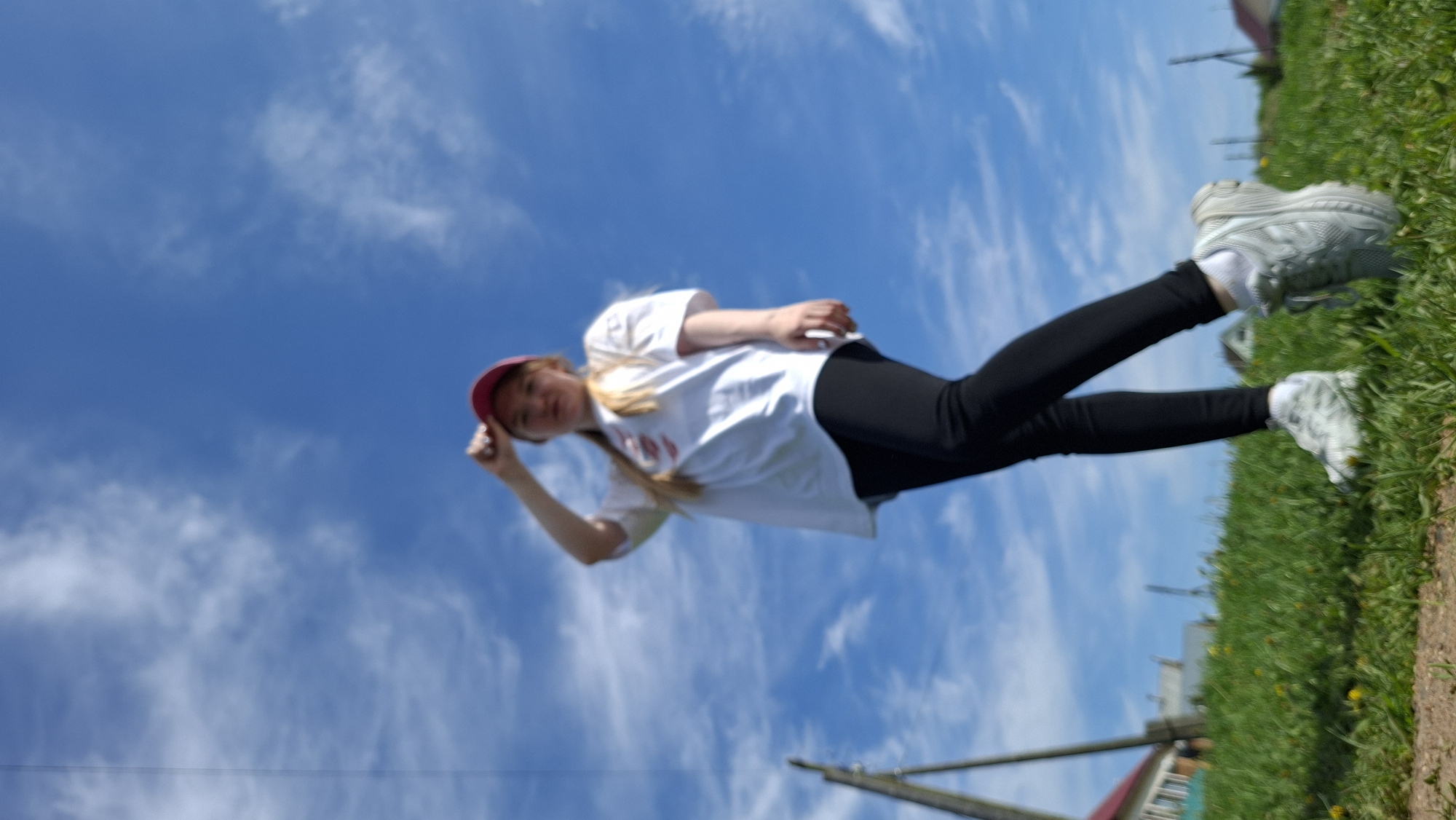 The image shows a young woman in a white t-shirt, black leggings, and sneakers standing on a grassy area under a bright blue sky with scattered clouds. She is wearing a red cap and appears to be adjusting or holding it with her hand. The background includes elements suggesting an outdoor setting, possibly a rural area or outskirts of a city, with signs of houses and vegetation. This is likely in Большой Букор, Russia, given the location information. The image is rotated counterclockwise.
