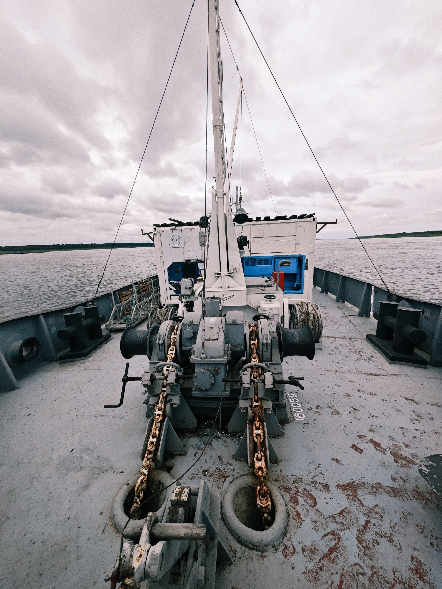 The image is a forward-facing view from the deck of a ship on a body of water, likely a river or lake, with a cloudy sky overhead. In the foreground, there are ship mechanisms, including a winch with a heavy chain extending downwards into a circular opening in the deck. The deck is grey and shows signs of wear and tear, with patches of rust and scuff marks. There are bollards on either side of the deck. In the mid-ground, there is a white structure with blue accents, possibly a cabin or equipment housing, with a ladder leading up to it. The water is calm and reflects the overcast sky. On the horizon, there is a faint green line of trees, indicating land. The image appears to have been taken in Zotino, Russia, though no specific landmarks or urban features are visible to confirm this. There are no people present in the image.