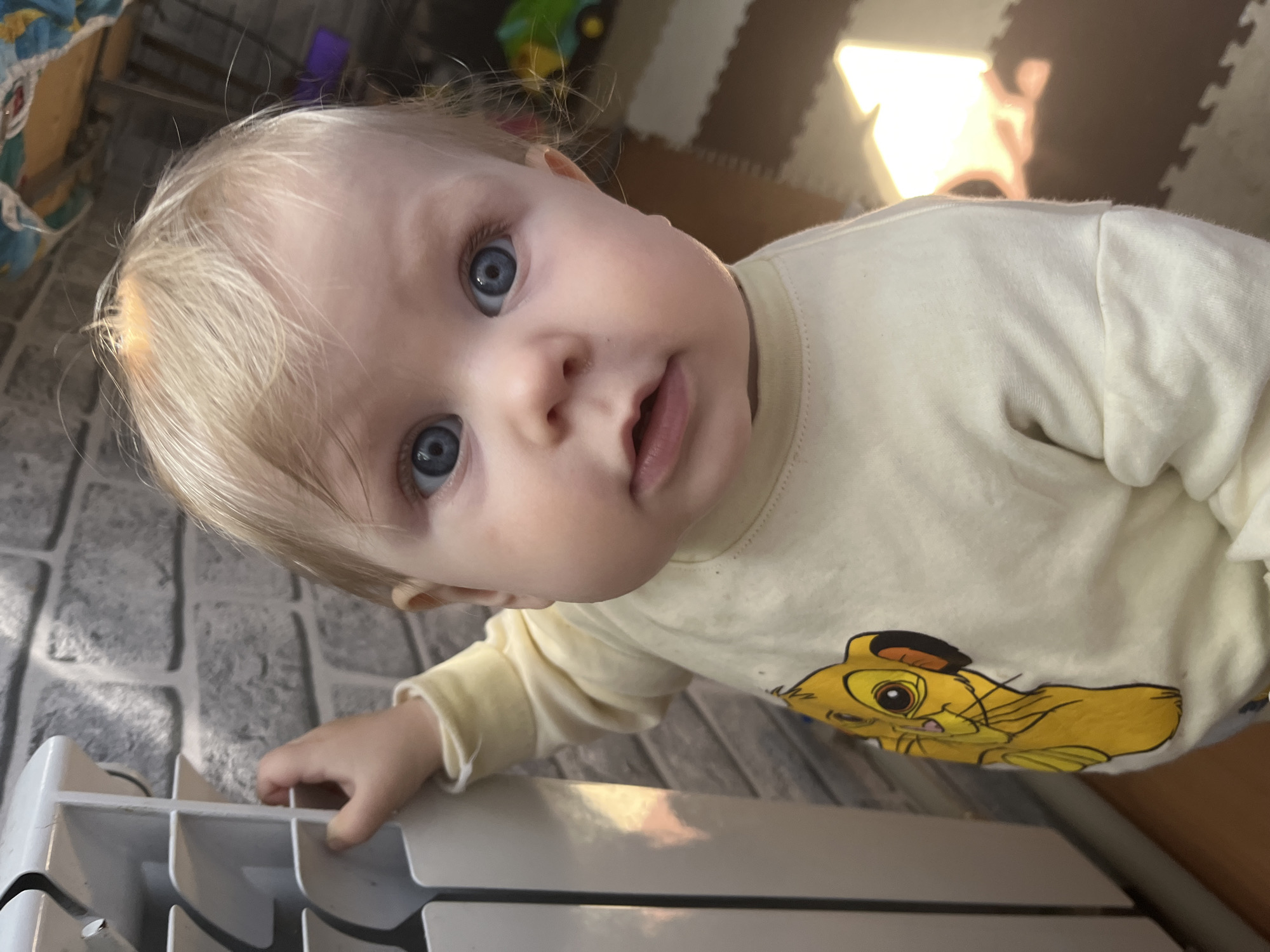 The image, captured from a slightly tilted perspective, features a baby or toddler looking directly at the camera with wide, striking blue eyes, fair skin, and light blonde hair. The child has a curious expression with a slightly open mouth. They are wearing a light yellow long-sleeved shirt adorned with a cartoon graphic of Simba from Disney's The Lion King on the chest.
In the immediate foreground, the baby's left hand rests on a white, slatted object, which appears to be a radiator or a heating unit. The indoor setting reveals varied flooring: a grey, textured surface resembling cobblestones or large tiles is visible on the left, transitioning to brown and lighter-toned interlocking foam play mats on the right and further into the background. A bright light source, likely natural sunlight, enters from the upper right, casting a warm glow and reflections within the scene.
The photograph conveys a candid, intimate moment of the child exploring their home environment during daylight hours in Vesyoloye, Russia. Beyond the character illustration on the shirt, no explicit text is visible in the image.