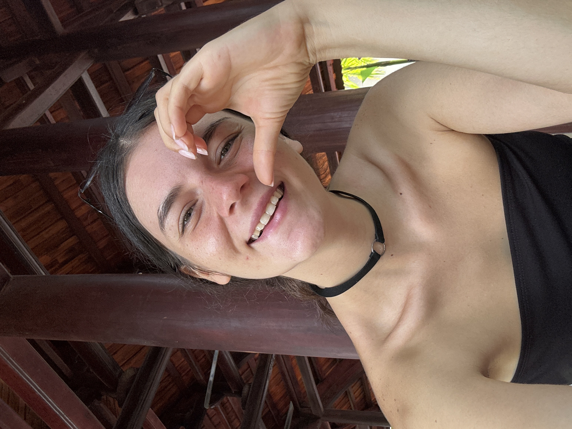 A young woman with dark hair and fair skin smiles directly at the camera, forming a heart shape with her right hand over her right eye. She is wearing a black strapless top and a black choker necklace. Her skin is adorned with freckles, and she has a slight tan. Behind her, a dark brown wooden pillar and ceiling structure are visible, suggesting an outdoor covered area. The overall scene appears to be within a resort or similar establishment in Mũi Né, Vietnam, as indicated by the tropical foliage visible in the background and the architectural style.