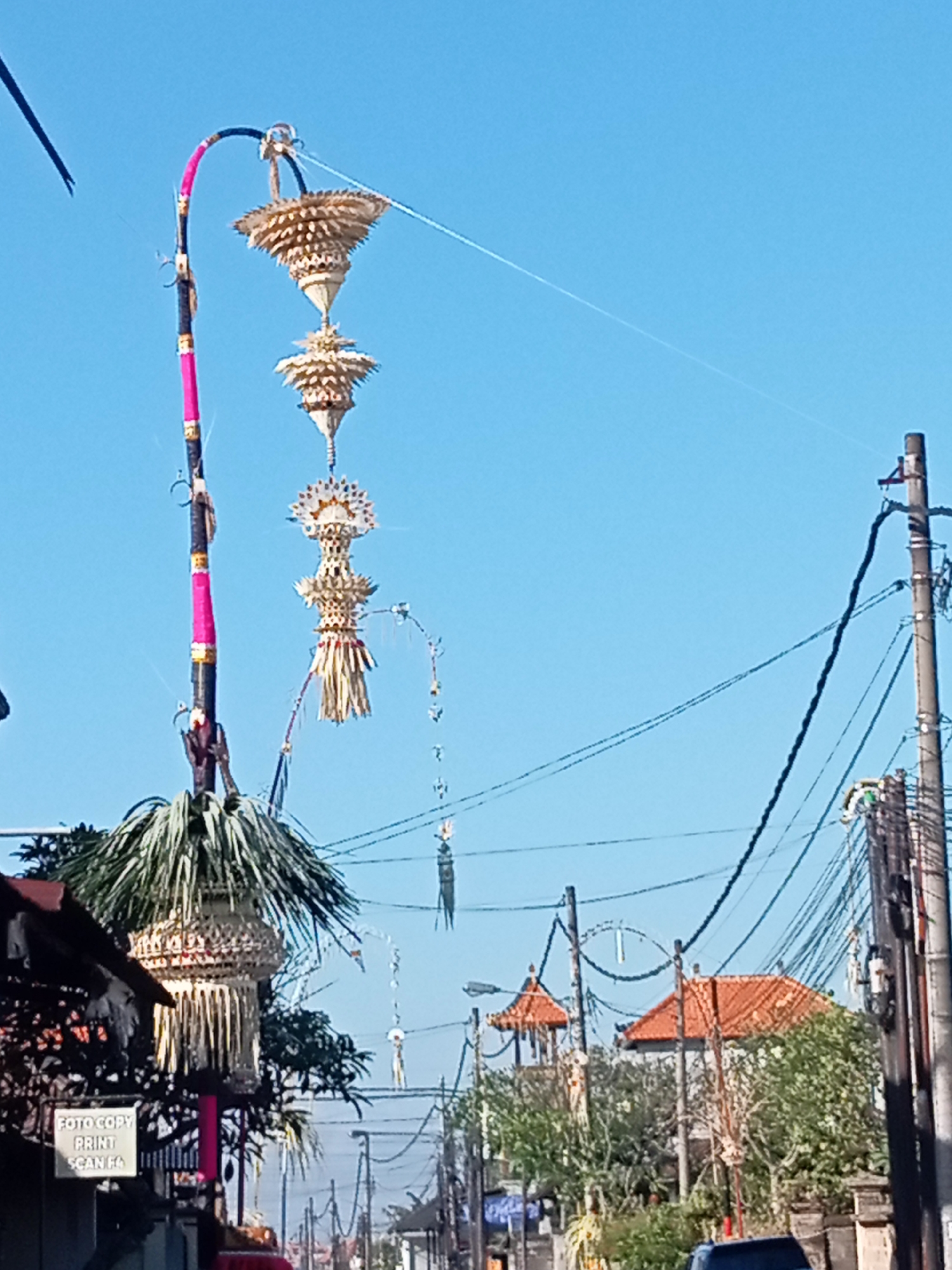 The image shows a street scene with traditional Balinese decorations. A tall, curved pole decorated with pink and black fabric and gold accents stands prominently on the left, adorned with several tiers of intricately woven, golden decorative elements. These elements appear to be made of natural materials like palm leaves and straw. Several strings extend from the top of the pole into the sky. In the background, there are more poles with similar decorations, as well as utility poles with a complex network of electrical wires. Buildings with orange tiled roofs are visible, along with trees and greenery. A sign that says "FOTO COPY PRINT SCAN EQ" is affixed to a building on the left. The sky is clear and blue. The scene depicts an outdoor setting, likely a street in a populated area, decorated for a festival or cultural event.