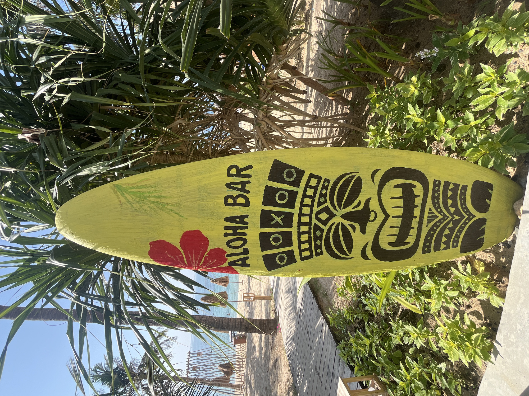 A bright yellow surfboard-shaped sign, painted with a red hibiscus flower and a black tiki face, stands upright on a sandy beach. The sign reads "ALOHA BAR" in black lettering. The tiki face features wide eyes, a prominent nose, and a gaping mouth with jagged teeth, all outlined in black. Above the tiki face, there is a checkered pattern with black and white circles and Xs. The surfboard is positioned in front of lush green foliage and the trunk of a palm tree, with its fronds extending across the top of the image.
In the background, a sandy beach stretches towards the turquoise ocean. Wooden planks form a pathway leading towards the water. Several straw umbrellas are spaced out on the sand, and a few beach chairs are visible. The sky is a clear, bright blue. The scene depicts a tropical beach bar in Nha Trang, Vietnam.