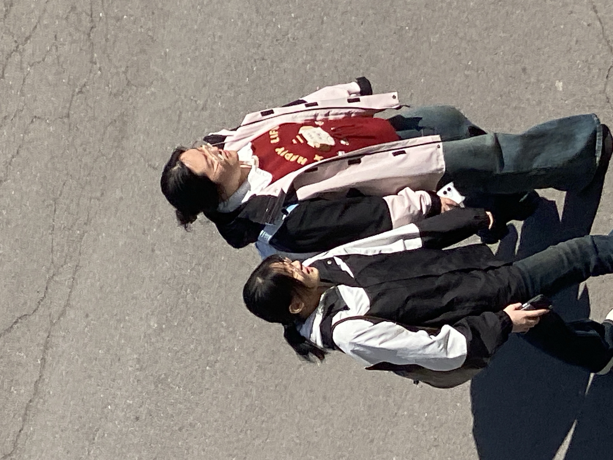 Two people are lying on a gray paved surface. The person on top wears a pink and white jacket over a red shirt with white lettering. The person below wears a black and white jacket and dark pants. They appear to be outdoors in daylight. The location is Zhongtun, China.