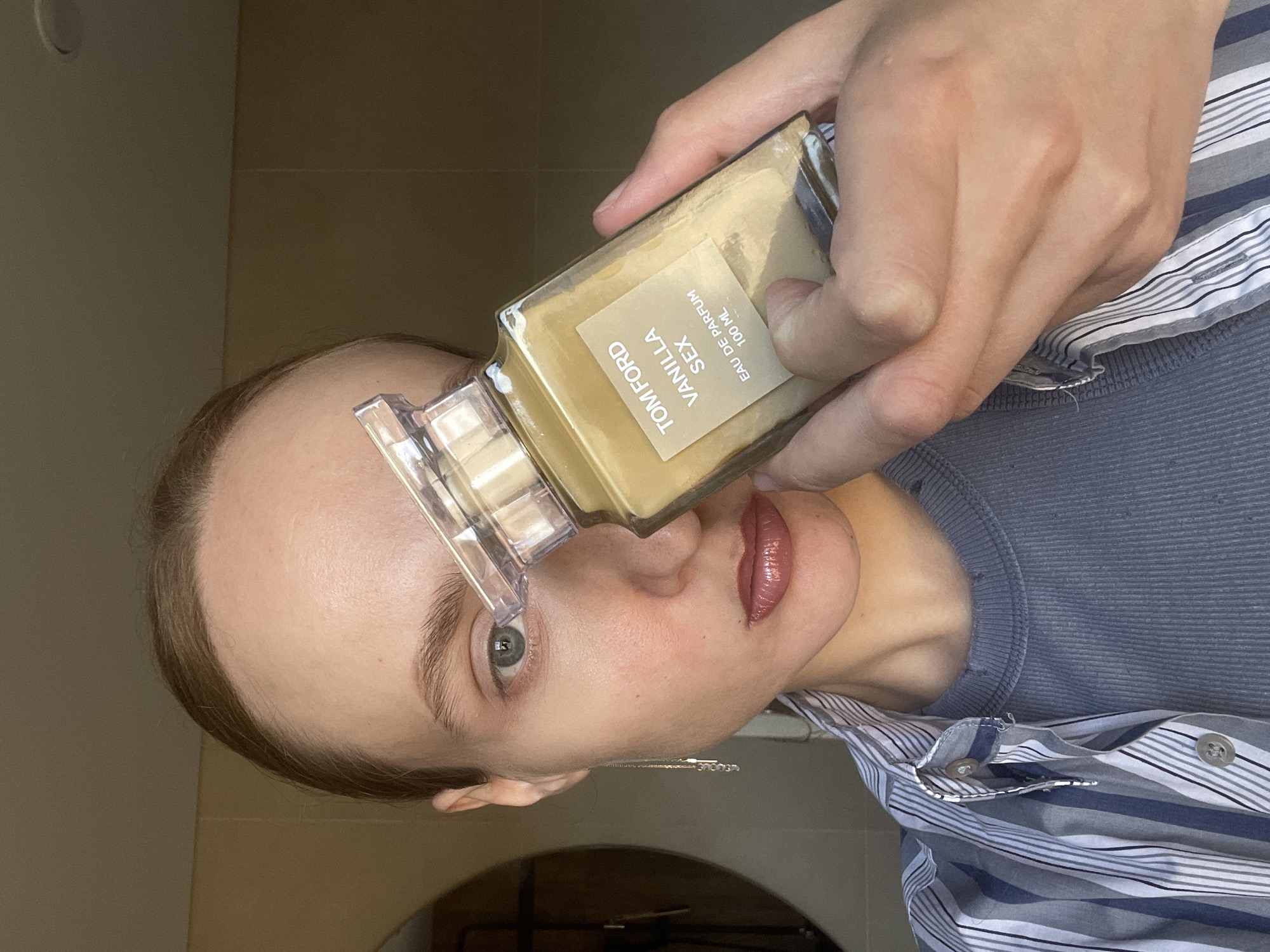 The image shows a person holding a bottle of Tom Ford Vanilla Sex Eau de Parfum in front of their face. The bottle is slightly tilted, with the label visible, indicating the brand and product name. The person's face is partially obscured by the bottle, but their eyes and lips are visible. The background appears to be an indoor setting, possibly a bathroom, with neutral-toned walls. The person is wearing a striped shirt and a solid-colored top underneath.