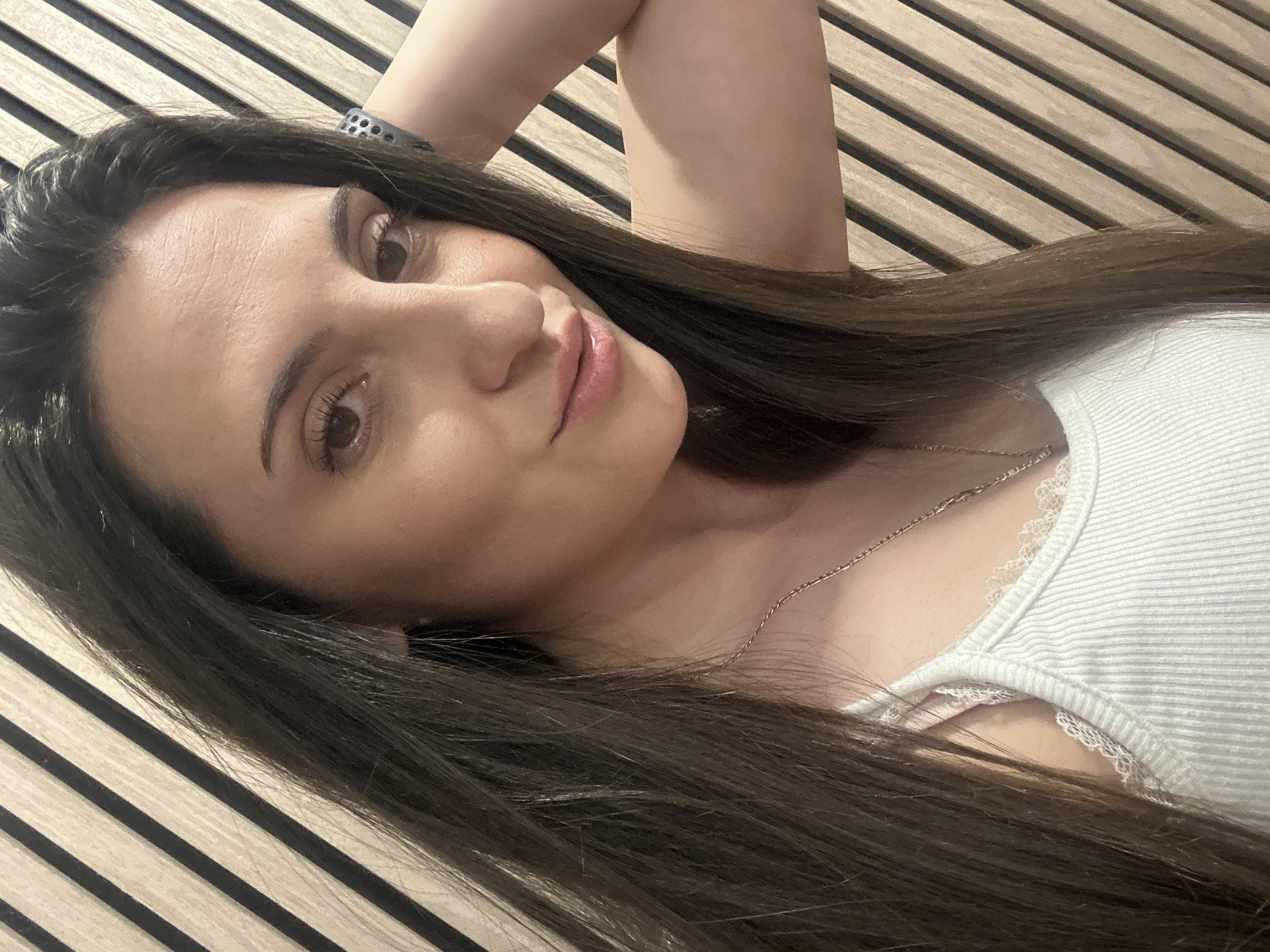 A woman with long, dark hair is lying down, facing the camera. She is wearing a white tank top with lace trim. A thin gold necklace is visible around her neck. She has brown eyes and light skin. Her left arm is bent and raised behind her head, with a watch on her wrist. She is lying on a surface with parallel wooden planks that have alternating light and dark tones. This image was not taken in Beliakovets, Bulgaria.