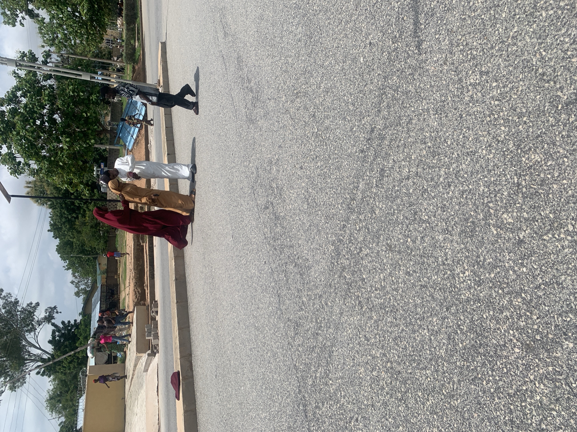The image shows a group of people on a street in Jalingo, Nigeria. The scene is captured from a slightly elevated perspective, looking down onto the asphalt road. In the foreground, three individuals are standing together. The person on the left is wearing a black and white patterned shirt and dark pants. Next to them, a person in a white robe is visible. To the right of the person in white, another individual is wearing a tan-colored robe. The fourth person, on the far right, is wearing a dark red robe.
Further in the background, along the side of the road, several other people are present, some appearing to be working or gathered. There are trees and buildings visible, suggesting an urban or semi-urban environment. The sky is overcast, indicating it might be a cloudy day. The overall atmosphere appears to be casual, with people going about their day. The lighting suggests it is daytime, likely mid-morning or afternoon. No text is visible in the image.