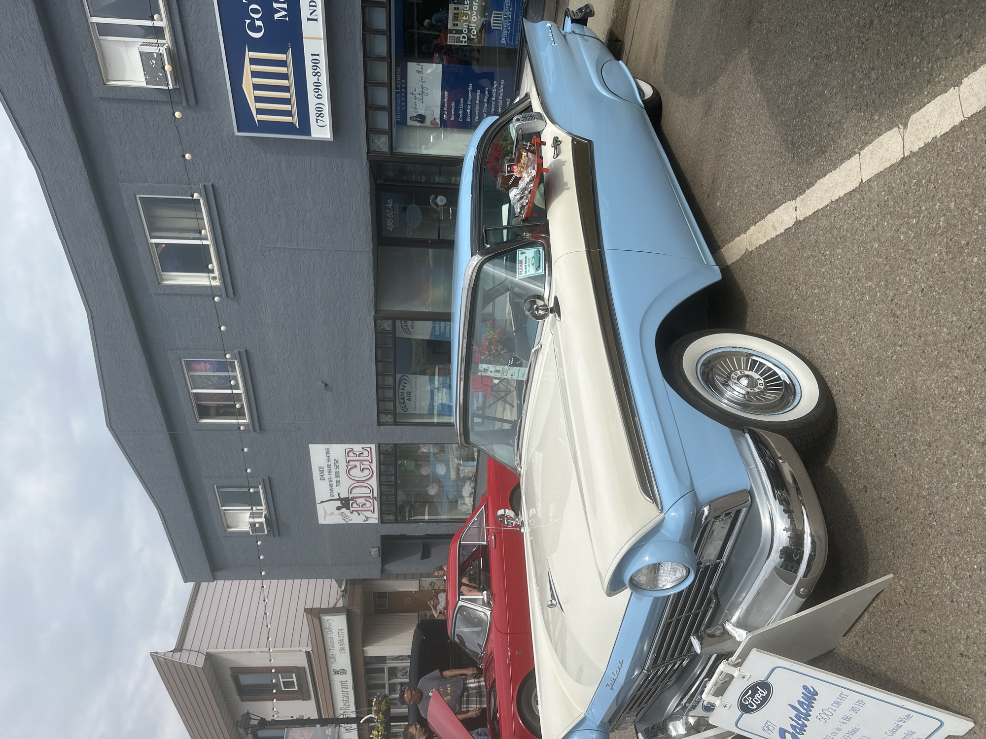 This image captures a classic car show event in Leduc, Canada, likely on a cloudy day. The primary focus is a two-tone blue and white 1957 Ford Fairlane 500 2-Door Hardtop, prominently displayed on a street. The car is in pristine condition, featuring whitewall tires and gleaming chrome accents. A sign in front of the car provides details about its specifications, including engine size, horsepower, and color.
In the background, a row of commercial buildings lines the street. One building, painted gray, features signs for "Dominion Lending Centres," "The Edge" (offering dance, gymnastics, and figure skating), and "Clean My Air." Another building to the right is identified as a restaurant with a "Buffet Takeout Delivery" service. The buildings suggest a downtown or main street setting.
Several people are visible, mostly near the cars, indicating they are attendees or participants in the event. One person is leaning over a red car parked next to the Ford Fairlane, and others are seen walking around. The overall atmosphere appears to be casual and community-oriented, typical of a local car show. The presence of street parking with painted lines and the building facades suggest an urban or town setting. No specific time of day can be definitively determined, but the lighting suggests daytime.