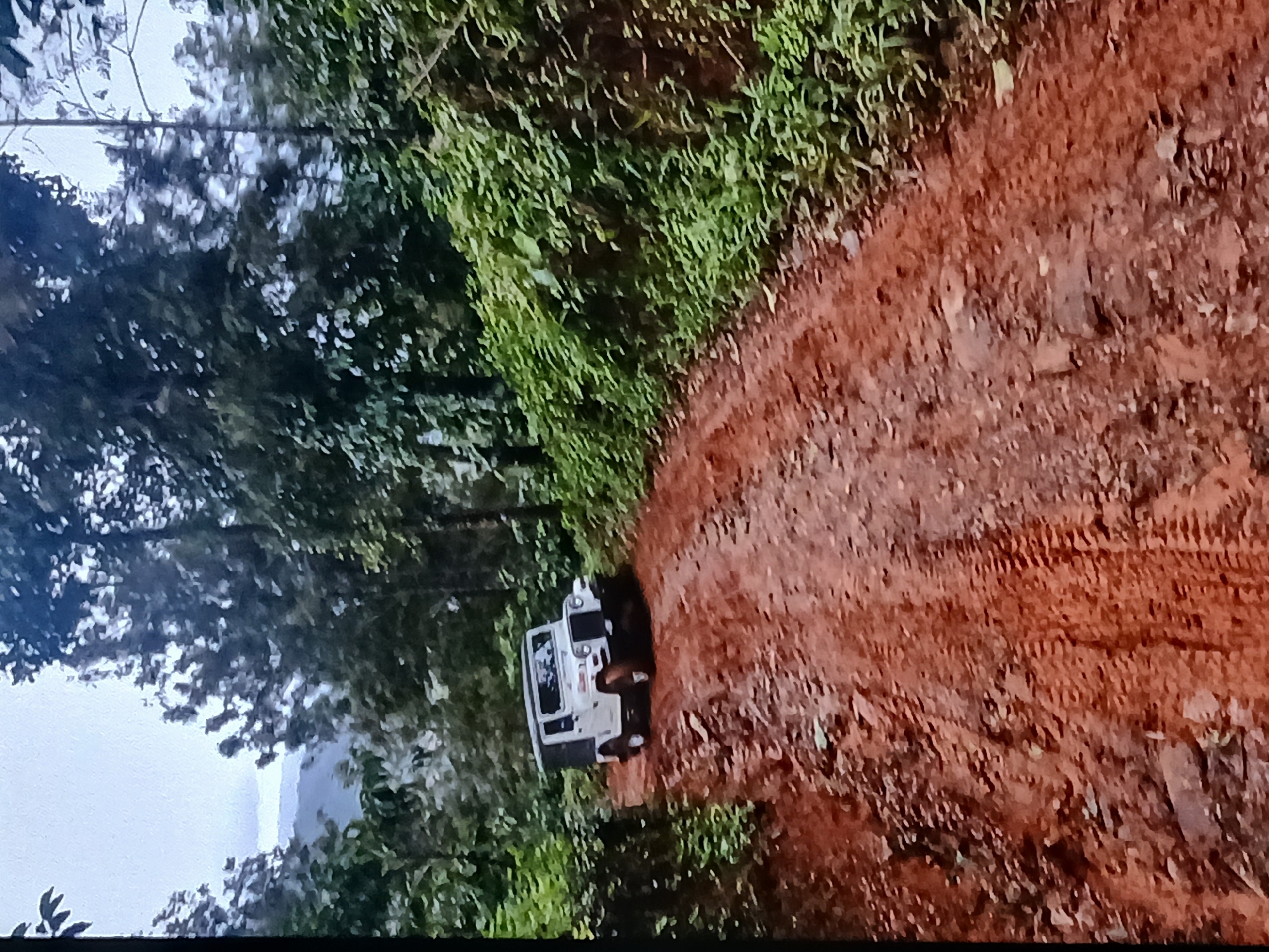 A white jeep navigates a mud-covered, tire-tracked path. Dense green foliage lines one side of the path. The photo was taken in a wooded area located near Bengaluru, India.
