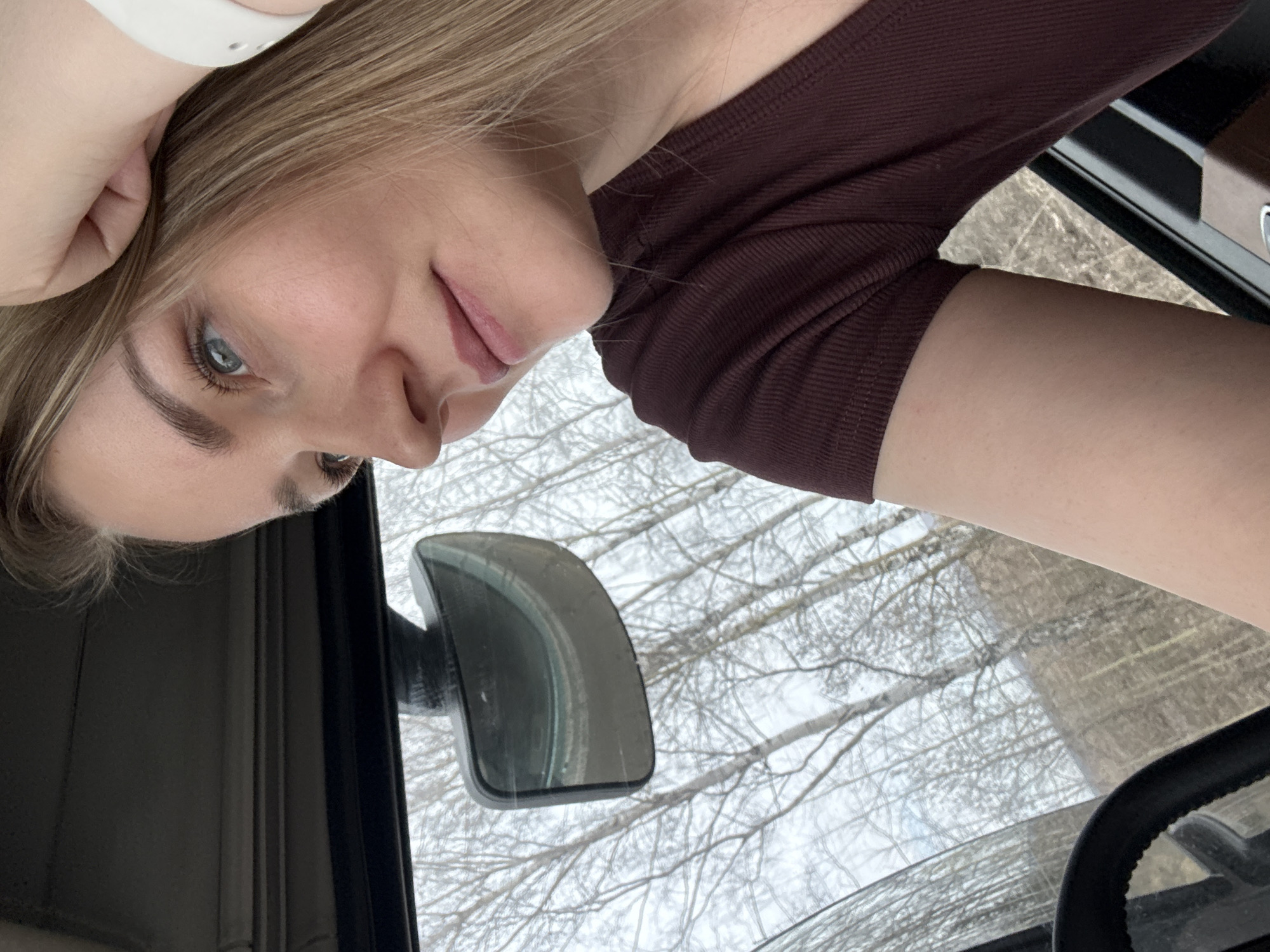 A woman with blonde hair, wearing a burgundy long-sleeved shirt and a white wrist device, is seated inside a vehicle. The vehicle's side mirror is visible, reflecting a view of trees with bare branches, likely a winter or early spring scene. The location cannot be determined as Kukui, Russia based on the image content alone. The steering wheel of the vehicle is partially visible in the lower right corner.
