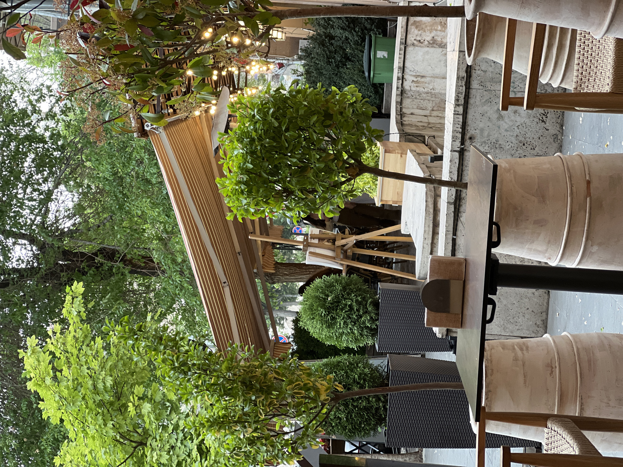 This is an outdoor dining scene, likely in Yerevan, Armenia. Tables and chairs are arranged on a patio, which is partially covered by a retractable awning. Lush greenery surrounds the dining area, including trees with small, leafy crowns and spherical bushes. One tree has strings of lights woven through its branches. Dining chairs are cylinder shaped and made of woven material. Two wicker outdoor chairs can be seen partially. In the background, there are other outdoor structures and a green trash bin. The atmosphere suggests a relaxed and inviting urban environment.