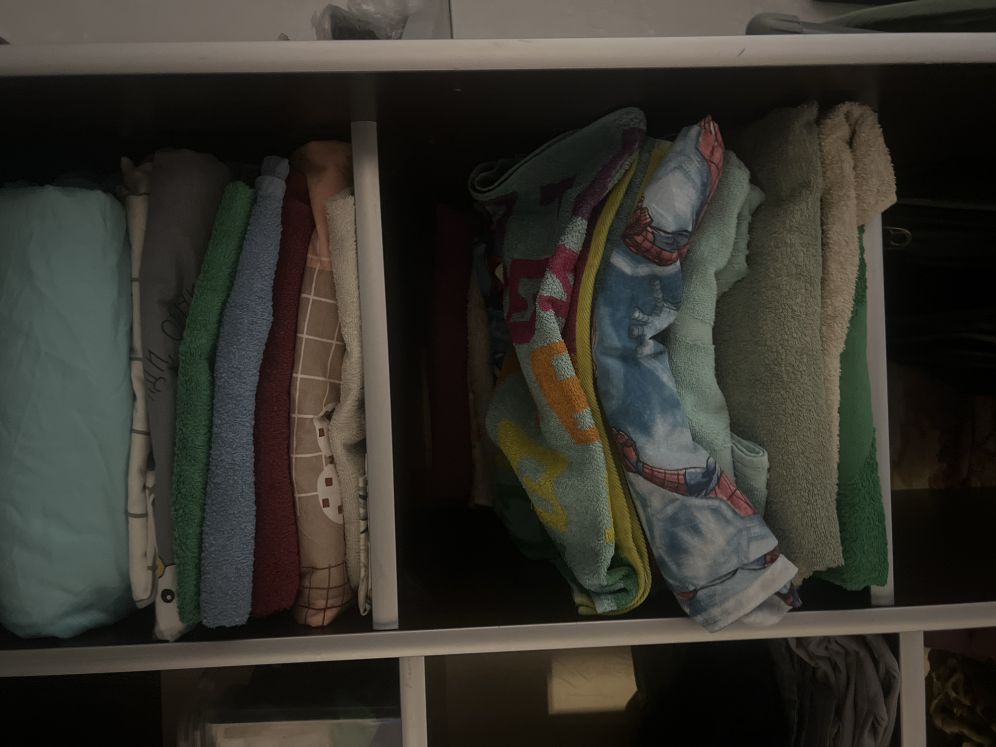 The image shows a close-up, eye-level view of shelves filled with neatly folded towels and bedding. The shelves are white and appear to be part of a closet or storage unit. The items are stacked from top to bottom. The top shelf has a large, light blue folded item, followed by a folded patterned fabric, a grey folded fabric, a green folded towel, a blue folded towel, a red folded towel, and a patterned folded fabric. The shelf below contains a stack of folded towels. The topmost towel on this shelf is teal with yellow and orange patterns. Below that is a blue towel with a Spiderman print. Underneath that is a light green towel, followed by a beige towel, and a green towel at the bottom. The background is dark. The image does not provide any information about the city of Surgut, Russia, or any people.