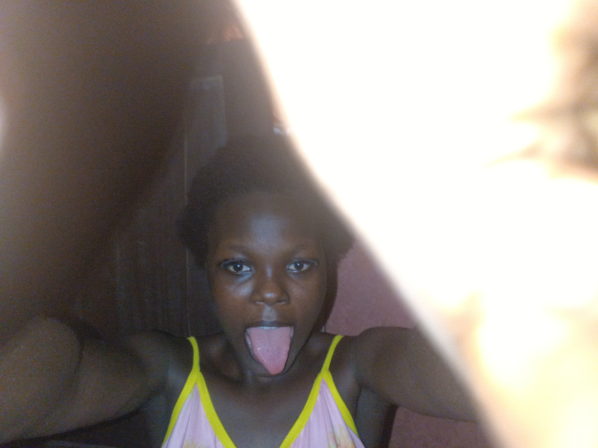 The image is a close-up portrait of a young individual with a dark skin tone and short, dark hair, captured in Onitsha, Nigeria. The subject faces the camera directly, mouth wide open, with their tongue fully extended downwards. They are wearing a light-colored (possibly pink or lavender) tank top featuring bright yellow shoulder straps. The scene is illuminated by an intense, likely artificial, light source (flash) originating from the camera's perspective. This light creates significant overexposure and a broad, bright white flare that dominates the right side of the frame, obscuring visual details. The background on the left is dark and indistinct, suggesting an interior setting with minimal ambient light.