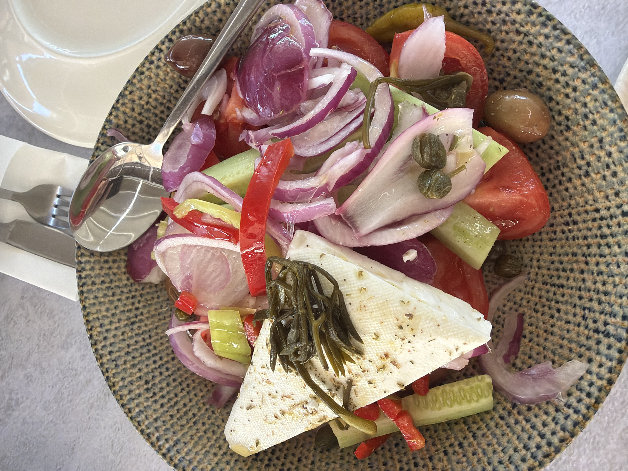 A close-up, overhead shot captures a Greek salad served in a textured, dark blue and tan bowl. The salad is a vibrant mix of ingredients, featuring a large, triangular wedge of white feta cheese seasoned with herbs. Interspersed are slices of red tomato, green cucumber, thinly sliced red onion, green capers, and two whole Kalamata olives. A portion of a shiny metal spoon rests on top of the salad, suggesting it's ready to be served or has been partially eaten. To the upper right, a white plate and silverware are partially visible, indicating a dining setting. The background is a light gray textured surface, possibly a table. The scene is located at a restaurant in Ofrynio Beach, Greece.