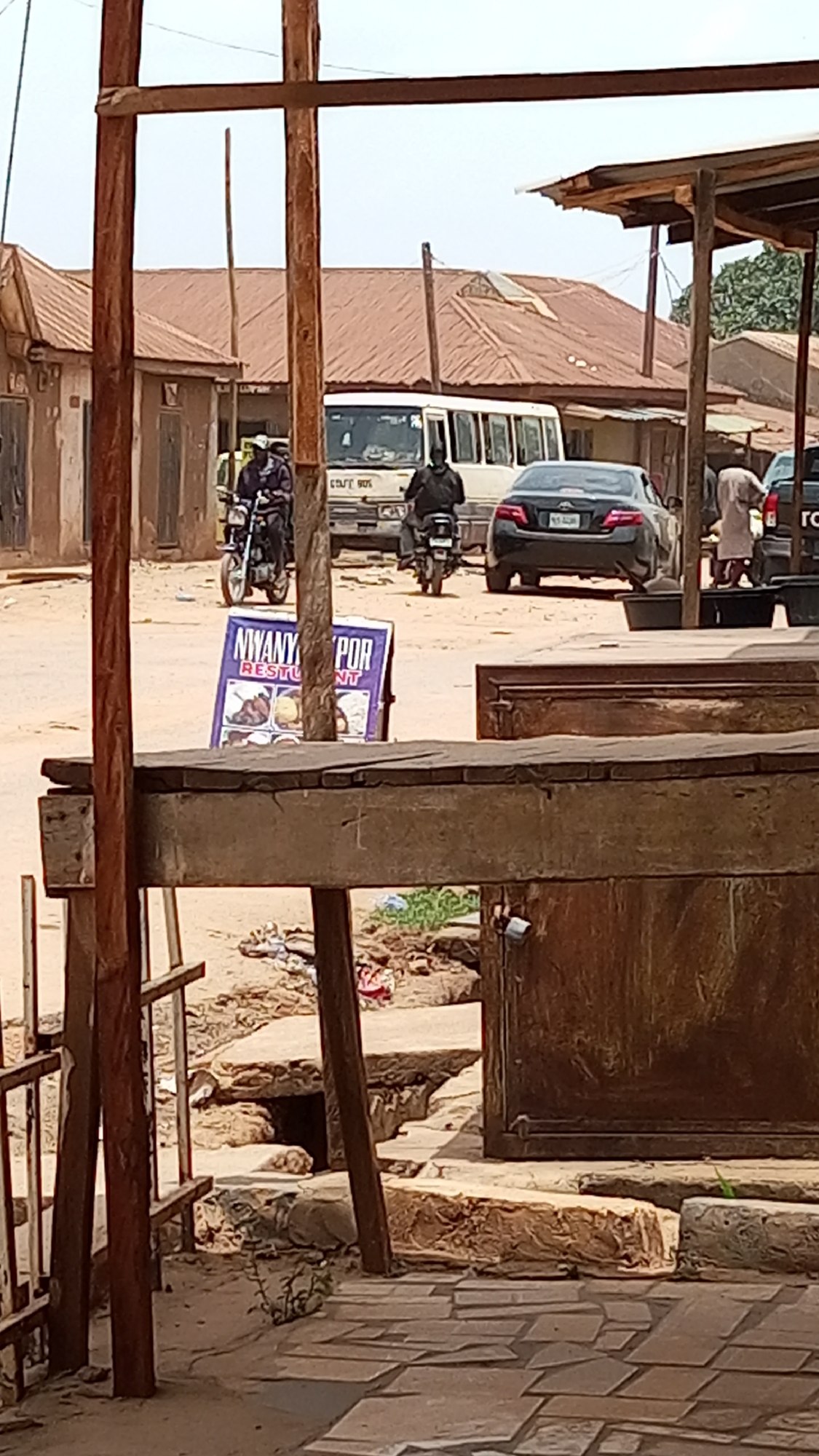 The image shows a street scene in Sabon Tasha, Nigeria. In the background, there is a white bus, a black car, and two motorcycles with riders. There are several buildings with brown roofs. In the midground, there is a sign for "Nyany Por Restaurant." In the foreground, there is a wooden structure. The ground appears to be dirt and stone. A person is standing on the right side of the frame.