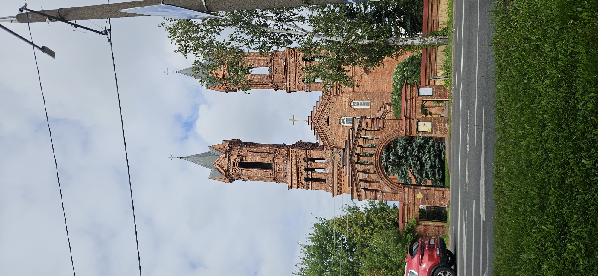 A low-angle, full-shot, vertical photograph captures a red brick church with two prominent bell towers against a cloudy sky. The church features a large arched entrance in the foreground, flanked by brick walls and smaller archways. A tall evergreen tree is visible through the main archway. To the left, a red SUV is parked on the side of a road, with a strip of green grass in the immediate foreground. Power lines stretch across the top of the frame, and a concrete utility pole with a street light and some wires stands on the right side. The church appears to be the Holy Assumption Cathedral in Vitsebsk, Belarus, indicated by its architectural style and the presence of crosses atop the towers. The scene is devoid of any people.