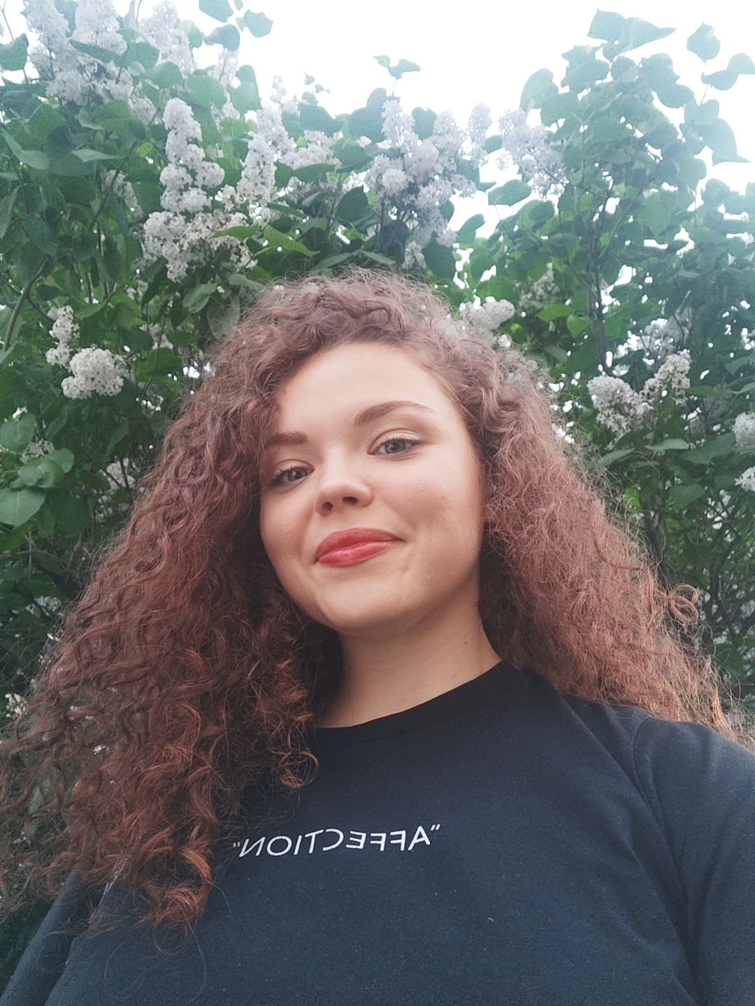 A young woman with auburn curly hair is posing for a selfie in front of a flowering tree. She is wearing a black sweatshirt with the word "AFFECTION" printed in reverse. The location is an urban setting, most likely in the city of Unknown, Unknown.