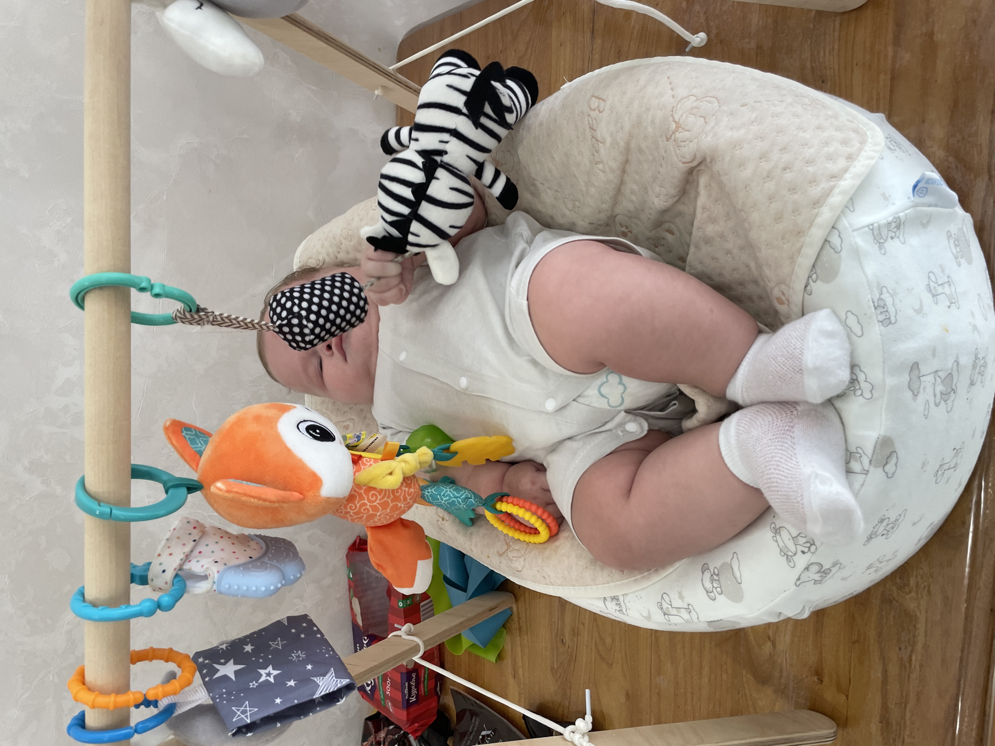 A baby is lying in a bouncer under a play gym. The baby is wearing a white onesie and white socks. The play gym has several toys hanging from it, including a fox plush toy, a black and white spotted rattle, and a zebra plush toy. The bouncer is light colored with a pattern of clouds and animals. The background shows a wall and a wooden floor. The image is taken indoors. There is no information available to determine the location in the city of Stavropol, Russia.