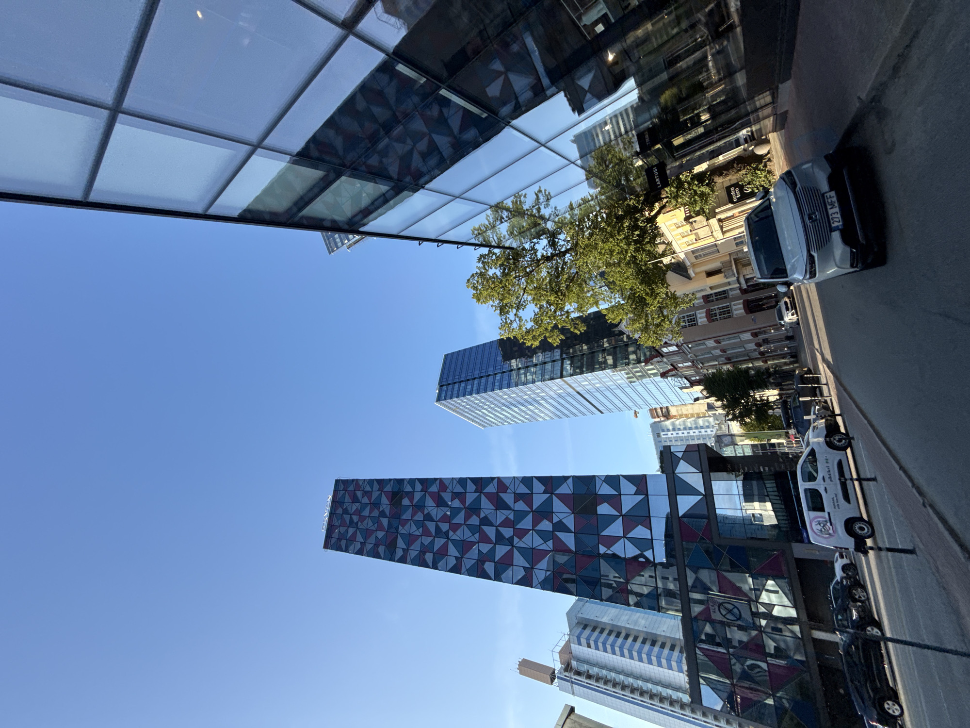 The image depicts a street scene in Tallinn, Estonia, with a clear blue sky overhead. Modern skyscrapers dominate the background, including a tall, slender building clad in a distinctive geometric pattern of red, blue, and white triangles. To its right, another skyscraper features a sleek, reflective glass facade. In the foreground, a street is lined with parked cars, including a white van with visible branding and a light-colored SUV with license plate "273 MFF". The street also has sidewalks with bollards and a tree providing some shade. The architecture suggests a contemporary urban environment with a mix of old and new buildings.
