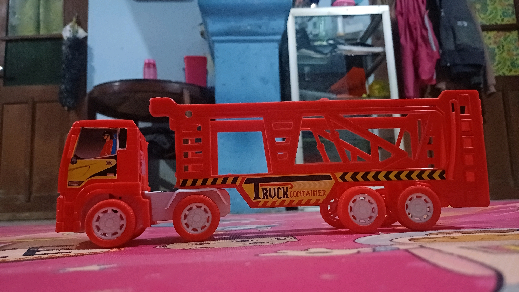 An orange toy truck with a blue and yellow "TRUCK CONTAINER" logo is pictured from a low angle, resting on a pink surface with a patterned background. The truck has a detailed cab with a driver figure visible through the windshield. The rear of the truck features a red frame structure with open sections. The background shows a blurry indoor scene with wooden doors, a blue wall, and furniture. There are no people other than the toy figure in the truck. The location in the city of Wonokerso, Indonesia, is not discernible from the image.
