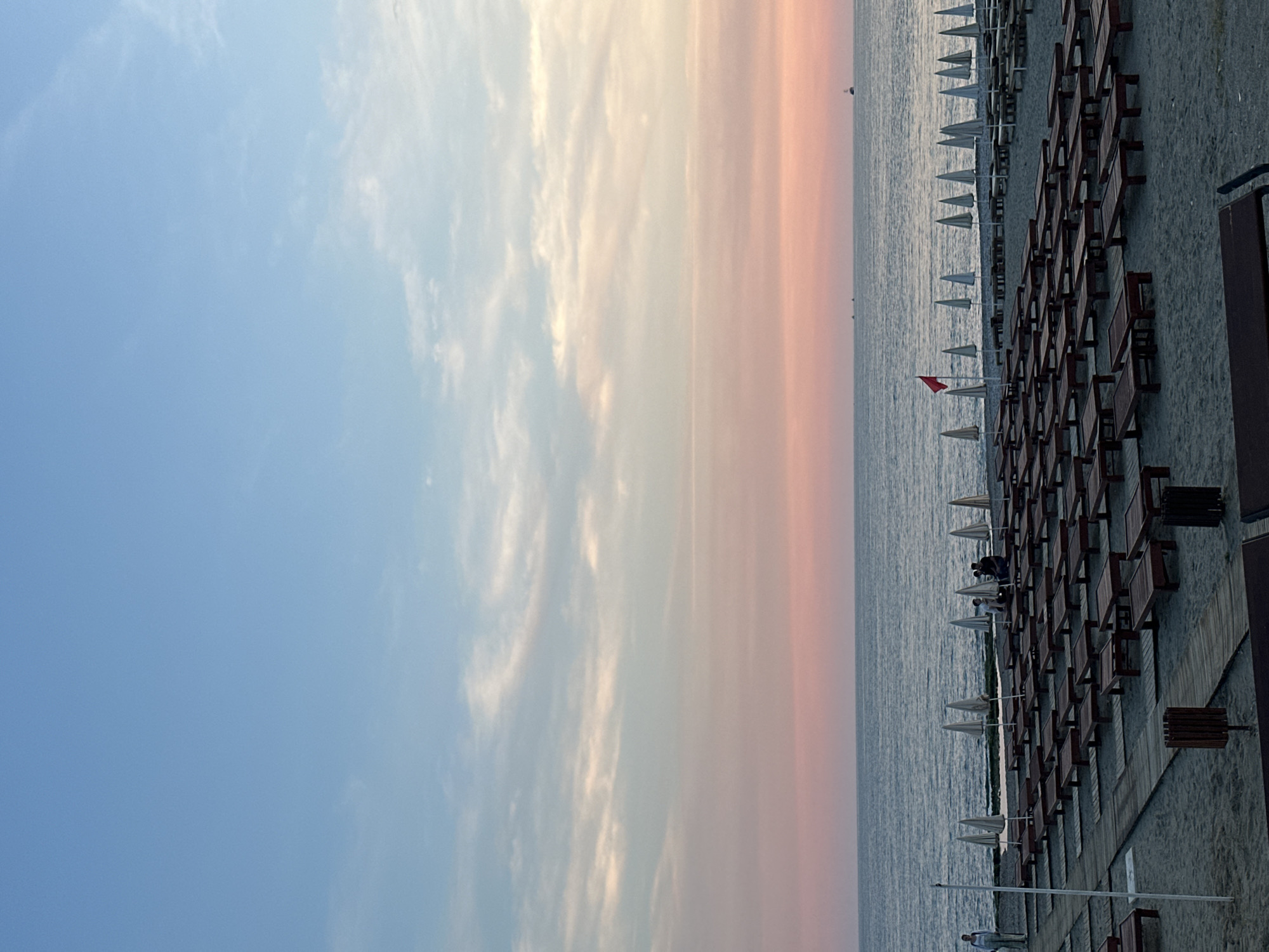 The image shows a beach scene in Alanya, Turkey at sunset. The sky transitions from blue to pink, with scattered clouds. The ocean is visible, with rows of white beach umbrellas and wooden beach chairs arranged on the sand. A red flag is placed on the beach. People are present on the beach between the umbrellas and chairs.