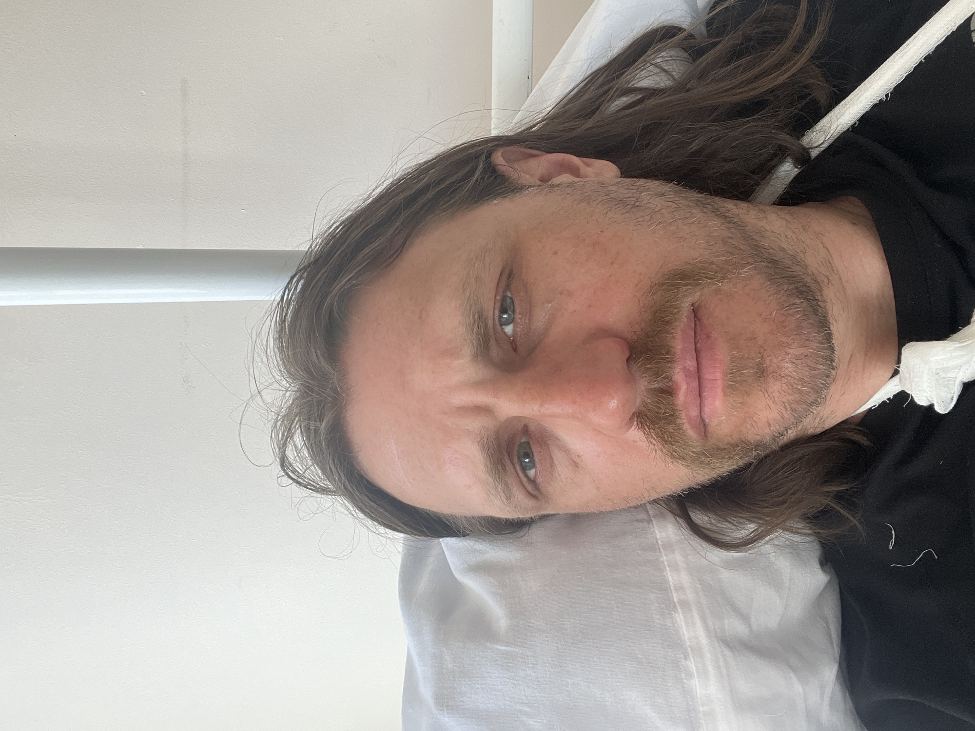 A man with long brown hair and a beard is lying on a white pillow. He is wearing a black hoodie. The location is Unknown, Unknown.