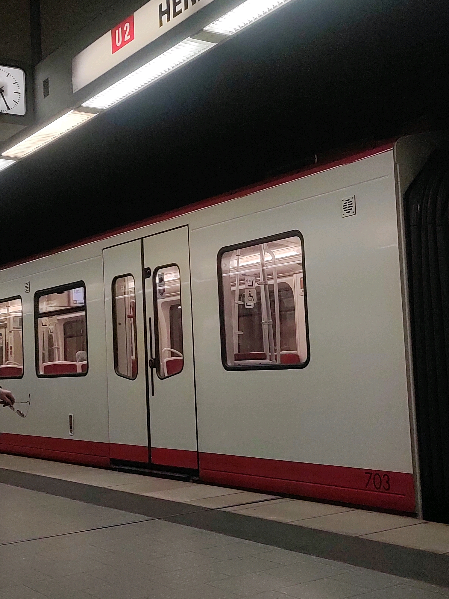The image shows a U-Bahn train at the Herderplatz station in Nuremberg, Germany. The train is white with a red stripe along the bottom and has the number "703" visible on its side. The station platform is made of gray tiles, and there is a clock on the wall showing approximately 12:05. The train doors are closed, and the windows offer a view of the interior, which includes red seats and handrails. There are signs indicating accessibility features within the train.