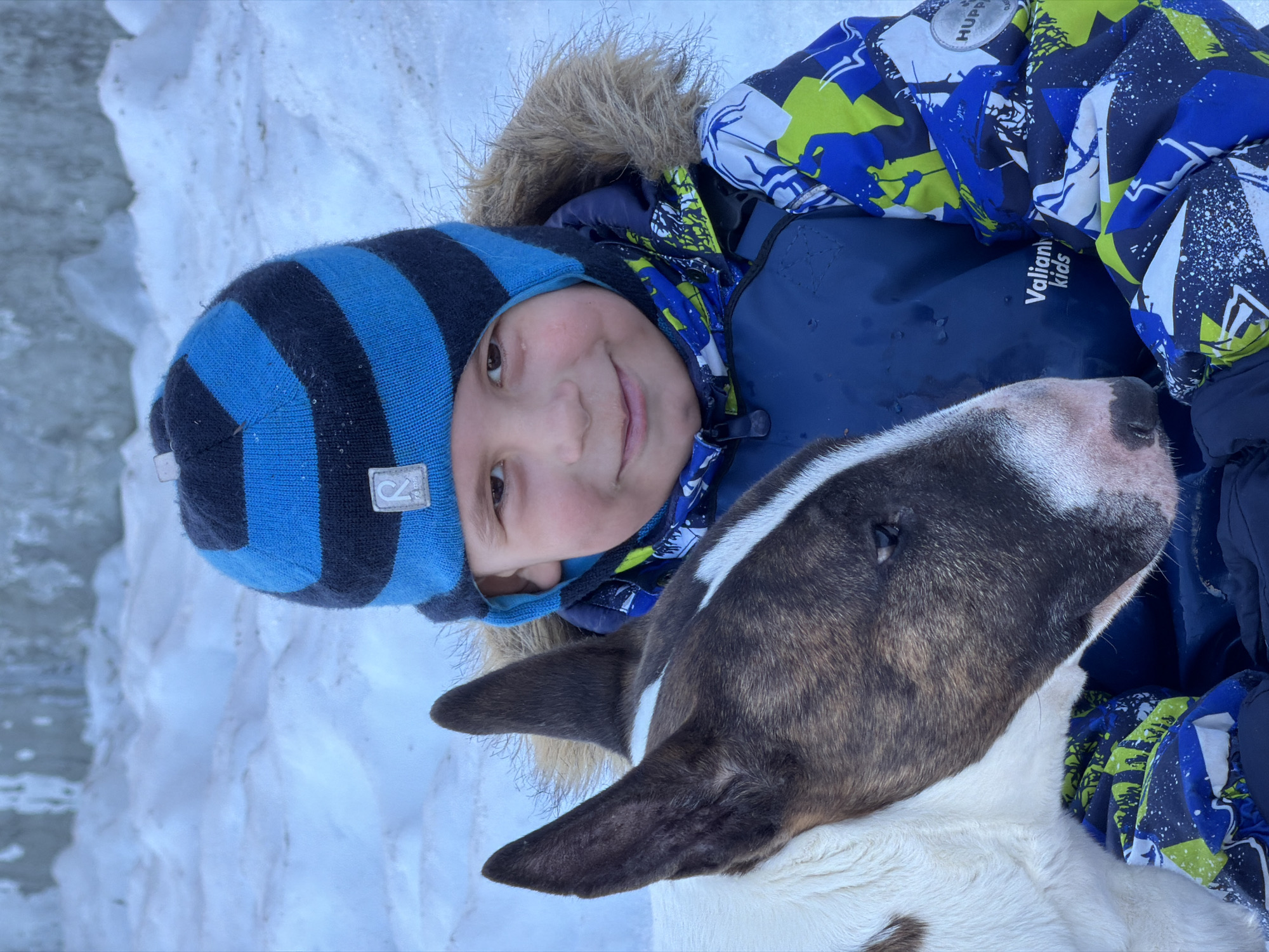 A young child, wearing a blue and black striped hat and a blue, green, and white patterned winter jacket, is lying in the snow with a bull terrier dog. The dog has a brown and white patterned head. The location appears to be outdoors in a snowy environment, possibly in the city of Chaikovsky, Russia. The child is looking towards the camera with a slight smile. The child is wearing a winter jacket with a fur-lined hood. There is snow visible in the background.