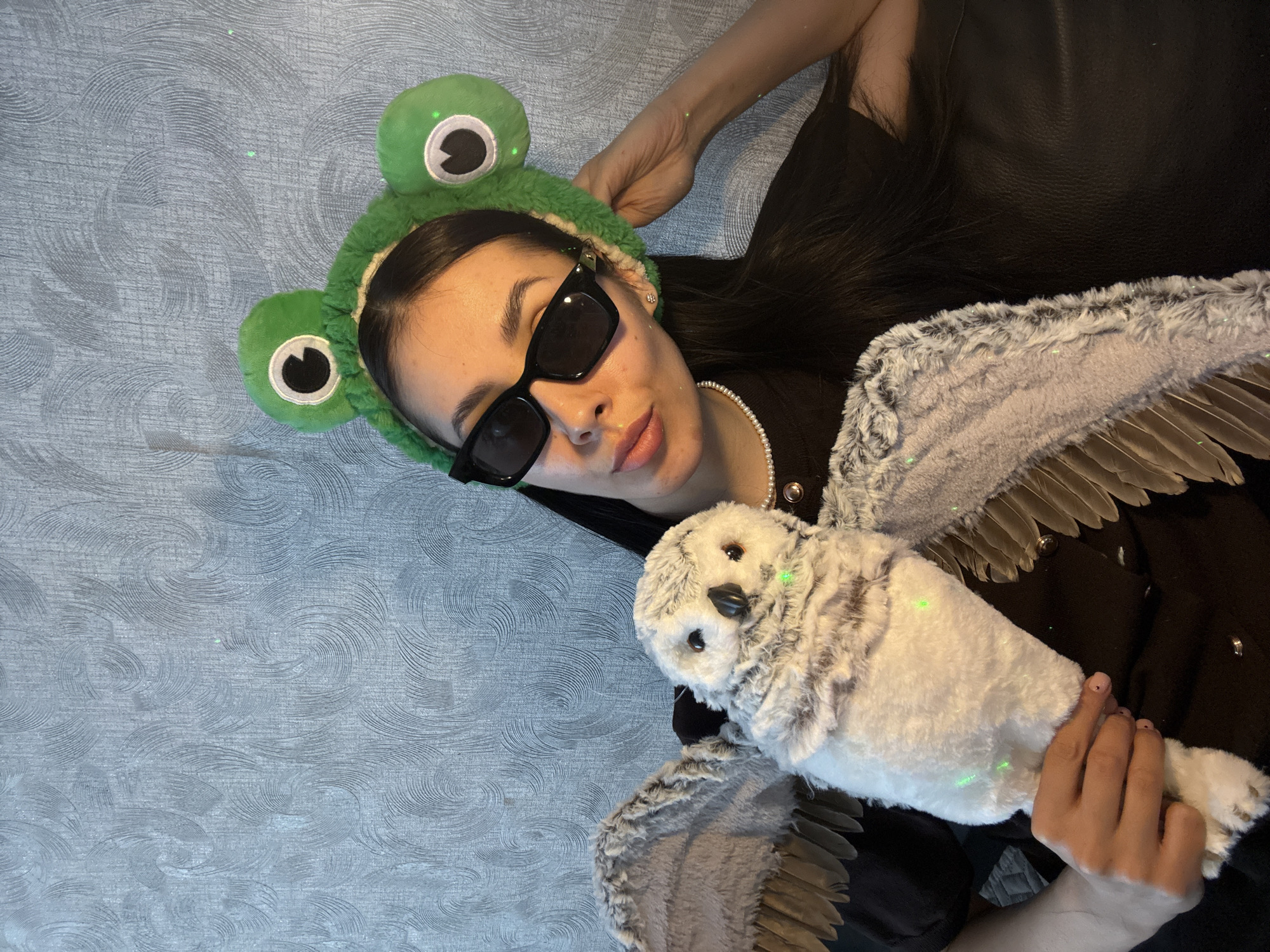 A person is lying on a surface with a textured pattern, possibly a rug or blanket. The person is wearing a black top, a pearl necklace, and sunglasses. They are also wearing a green headband with frog eyes attached. The person is holding a stuffed owl with wings spread out. The image contains green light dots that seem to be an element from a laser. The background seems to be a leather sofa. There is no information available to determine if this image was taken in Votkinsk, Russia.