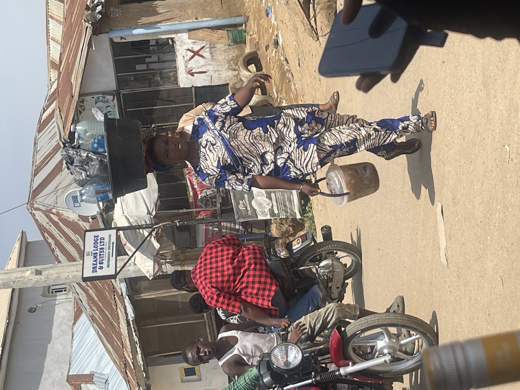 In a street scene in Abuja, Nigeria, a woman walks carrying a wide basin filled with plastic bottles of water and plastic bags on her head. She is wearing a patterned blue and white traditional outfit. In her right hand, she carries a clear plastic bucket containing food. To her left, a young man in a white tank top stands next to a red motorcycle. Behind him, another man in a red and black plaid shirt rides on the back of a motorcycle. In the background, buildings with corrugated metal roofs are visible, along with signs for "DREAMS LODGE & SUITES LTD" and a street vendor's stall. The ground is dusty earth. The overall atmosphere appears to be that of a busy marketplace or thoroughfare.