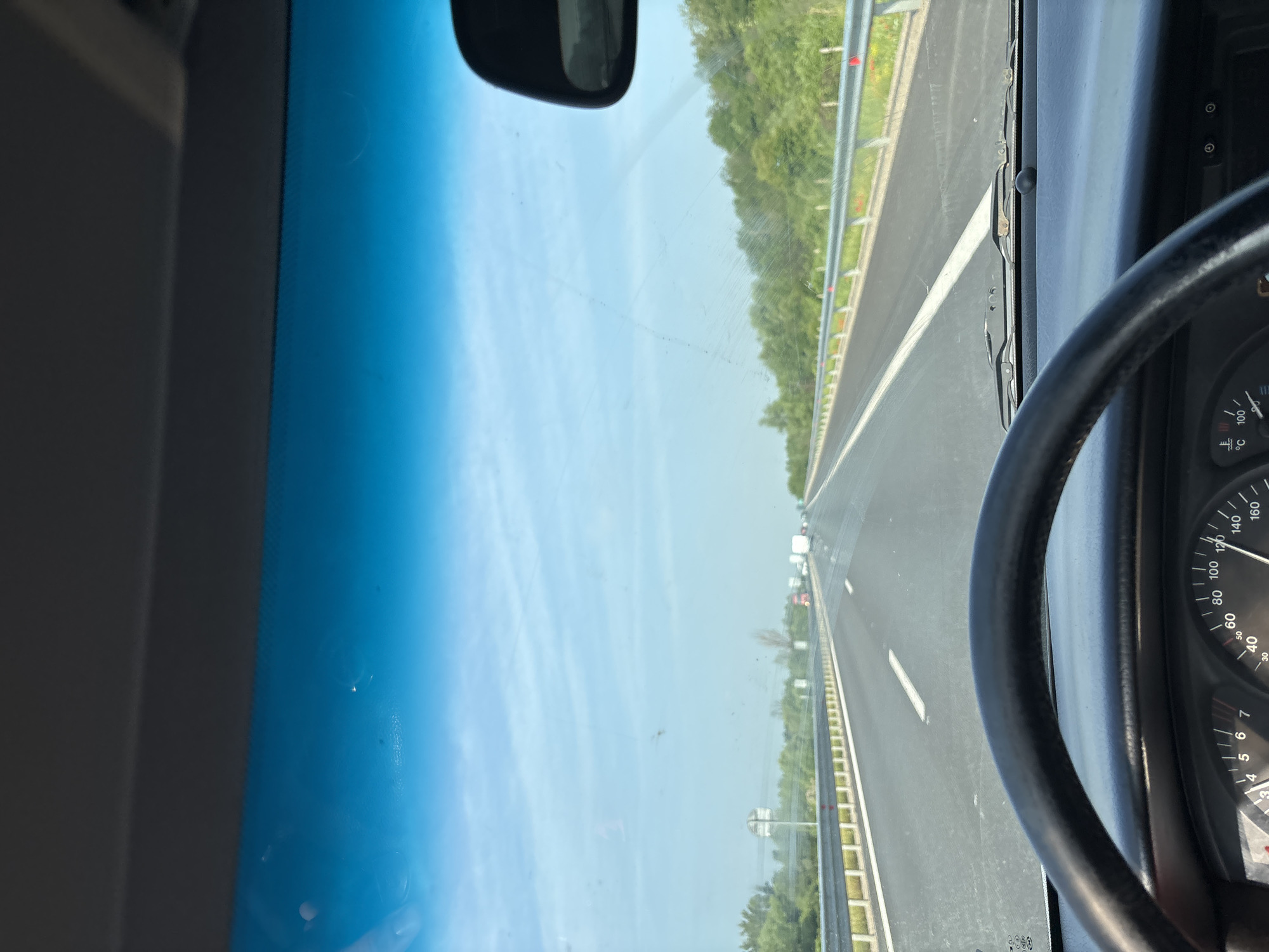 The image depicts a first-person perspective from the driver's seat of a vehicle traveling on a multi-lane highway in the vicinity of Dolni Bogrov, Bulgaria. The road surface is asphalt, with clear white lane markings visible, including a solid white line on the left and a dashed line on the right, indicating movement in the rightmost lane. Metal guardrails line both sides of the highway, beyond which extensive green vegetation, primarily trees and bushes, is visible under a bright blue sky with scattered light clouds. In the distance, other vehicles, including at least one truck, are visible in the right lane, traveling in the same direction.
In the foreground, the top of a black, leather-wrapped steering wheel is partially visible at the bottom-left. Below the windshield, a dashboard is present, with a portion of the instrument cluster visible at the bottom. A speedometer indicates a speed of approximately 120 km/h, and a temperature gauge is also discernable. The car's windshield displays various smudges, dust particles, and streaks consistent with wiper blade action. The upper portion of the windshield features a blue tint. Part of the interior rearview mirror is visible on the right side of the frame. The base of the windshield wipers is visible just above the dashboard. The bottom edge of a gray sun visor is visible at the very top of the image.