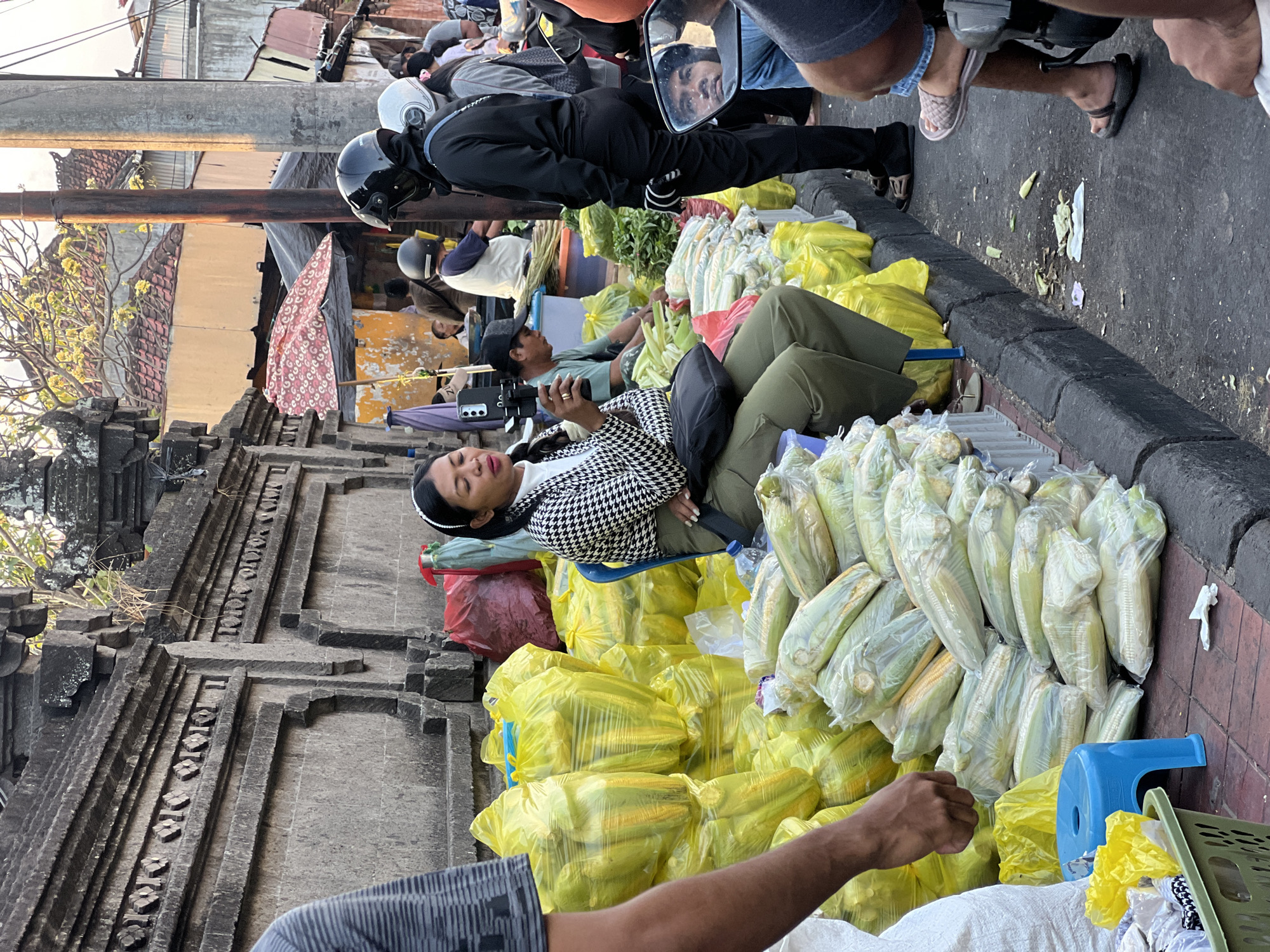 In Denpasar, Indonesia, a street vendor sits on a blue chair in front of a large display of corn packaged in yellow plastic bags. She wears a black and white houndstooth jacket, green pants, and a white headband. She holds a smartphone on a small tripod, seemingly recording. Other vendors and shoppers are visible in the background, some with helmets and on motorcycles, suggesting a bustling market atmosphere. The scene is set against a backdrop of a stone wall with ornate carvings and a variety of market stalls and buildings. The ground is paved with bricks and asphalt, littered with some discarded food waste.