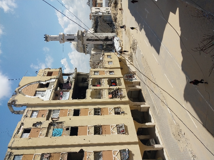 The image depicts a scene of extensive urban damage in Khan Yunis, Palestinian Territory, under a clear blue sky with scattered white clouds. In the foreground and left, a multi-story, light-colored apartment building stands significantly damaged. Its upper floors show severe structural compromise, with large sections of the facade missing, exposed interiors, and numerous shattered windows. Smaller pockmarks are visible across its surface. Laundry, including a blue tarp and various clothes, hangs from several damaged windows and balconies. The ground floor features recessed arched openings, with one parked vehicle partially visible within an opening.
A wide, sandy road with partially obscured white lane markings occupies the mid-ground. Debris and scattered rubble are visible along its edges. Overhead electrical or communication wires crisscross the frame, casting distinct shadows onto the road. Several individuals are present on the street: one person in dark clothing walks from left to right in the immediate foreground, largely in shadow. Further down the street, approximately five to six additional individuals are discernible, walking on the sandy road or amidst scattered debris.
In the background, to the right of the damaged apartment building, stands a prominent mosque with a white minaret and dome, also exhibiting significant structural damage, including missing sections of its walls. Beyond the mosque, extensive piles of rubble and the remains of other destroyed buildings indicate widespread devastation.