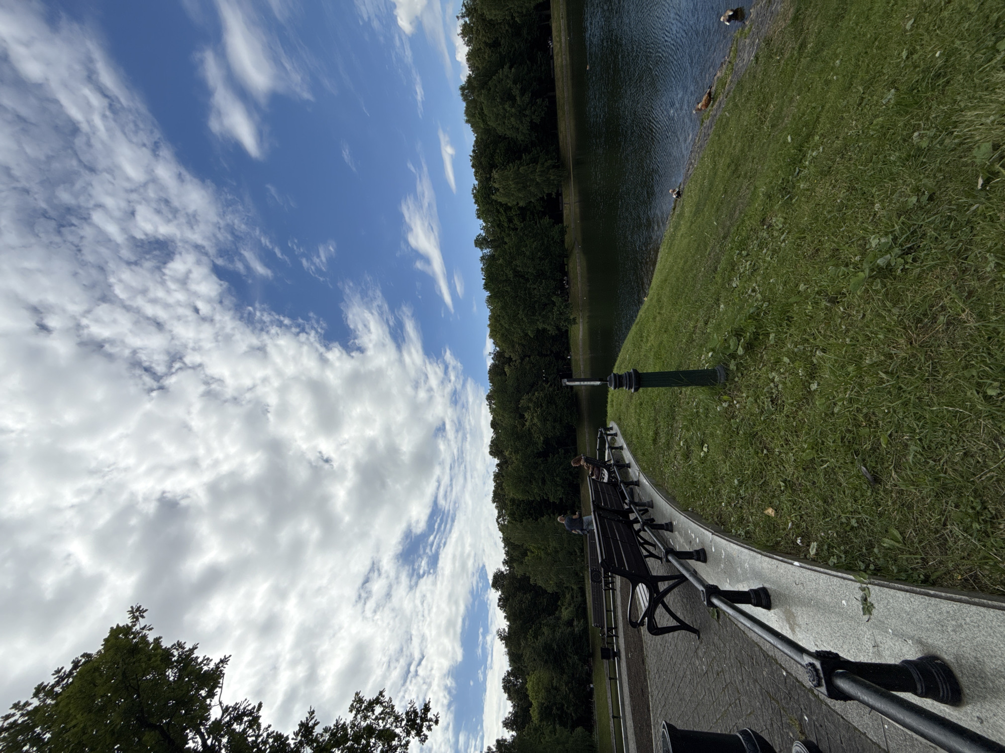 The image depicts a scenic view of a park in Moscow, Russia, likely during daylight hours given the bright sky and visible shadows. The foreground features a paved walkway with several empty park benches situated along a curved stone embankment. A black metal railing runs along the embankment. Beyond the walkway and embankment, a large body of water, possibly a lake or river, occupies the middle ground, reflecting the sky. Dense green trees line the far bank of the water, forming a backdrop of natural foliage. The sky above is a mixture of bright blue and scattered white clouds. In the water, there are a few ducks visible. One person is seated on a bench, and another person stands near a bench.