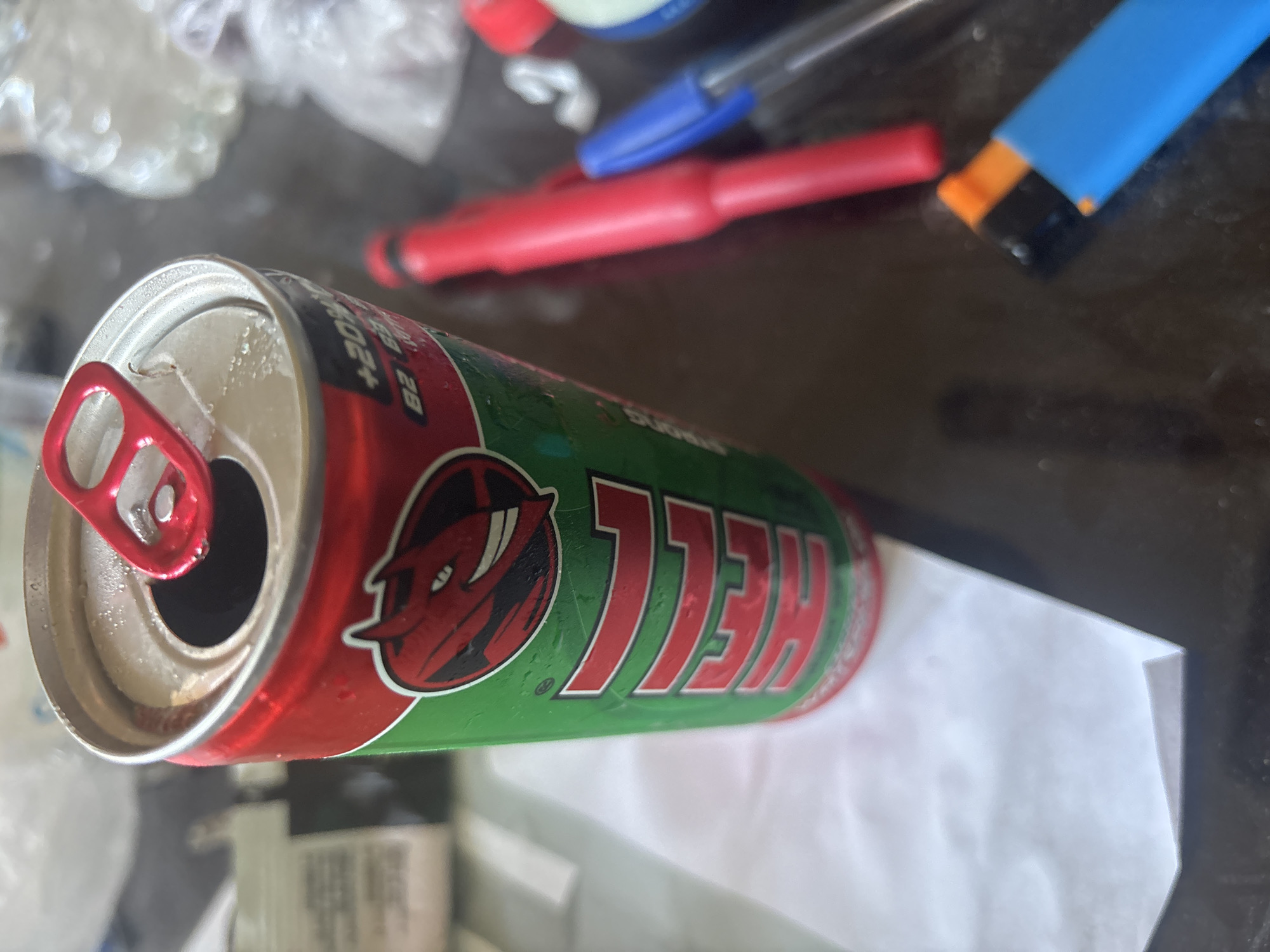 A close-up, slightly angled, eye-level shot shows a partially consumed energy drink can with a red pull-tab. The can is predominantly green and red with a cartoon devil logo and text, some of which is obscured. The can appears to be sweating, indicating it is cold. In the background, out of focus, are various objects including pens, a lighter, and what appear to be plastic bags. The surface the objects are on is dark and reflective, possibly a table. The image is taken indoors, in what appears to be a home or office environment, and there is no indication of the specific location in Sofia, Bulgaria.
