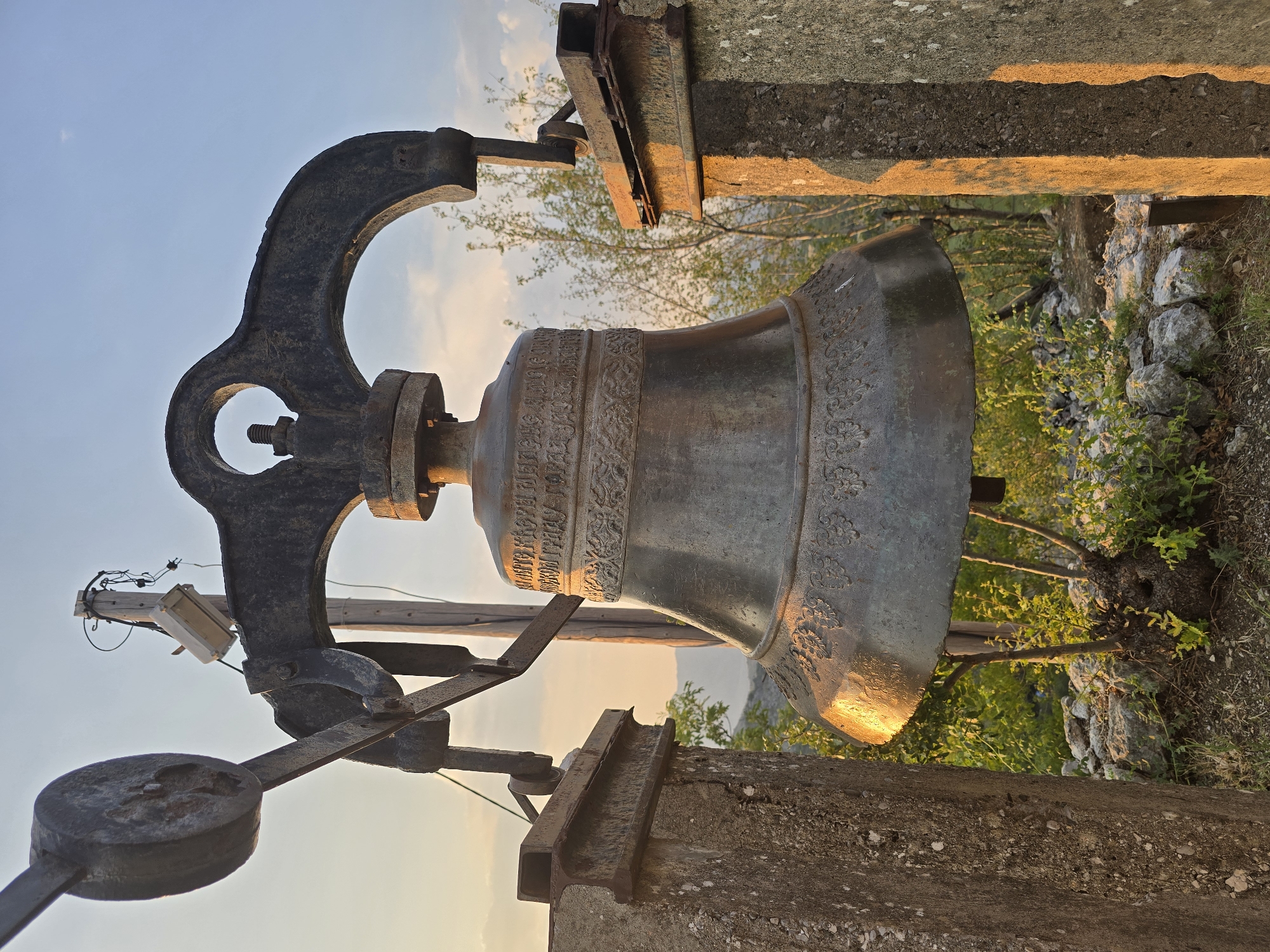 An old, weathered bronze bell hangs from a rusty metal yoke, which is supported by two concrete pillars. The bell has decorative bands around its rim and some visible inscriptions. It is mounted outdoors, with greenery and rocky ground visible behind it. In the background, hills are faintly visible, and the sky is a clear blue with some wispy clouds. The setting sun casts a warm, golden light on the scene, highlighting the textures of the bell and the concrete pillars. A wooden pole with electrical wires and a small box is visible behind the bell. The location is the town of Gornja Vrbica, Montenegro.